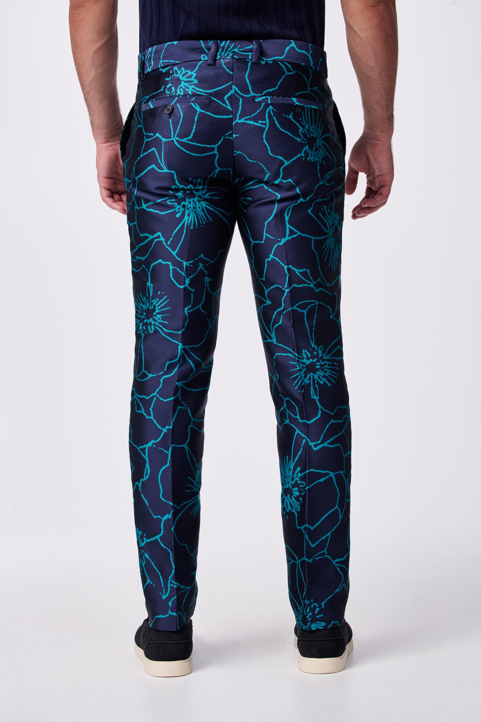A person is shown from the back wearing the Clyde Slim Trouser, featuring a dark blue base with a bold turquoise floral pattern, paired with black shoes. The background is plain white.
