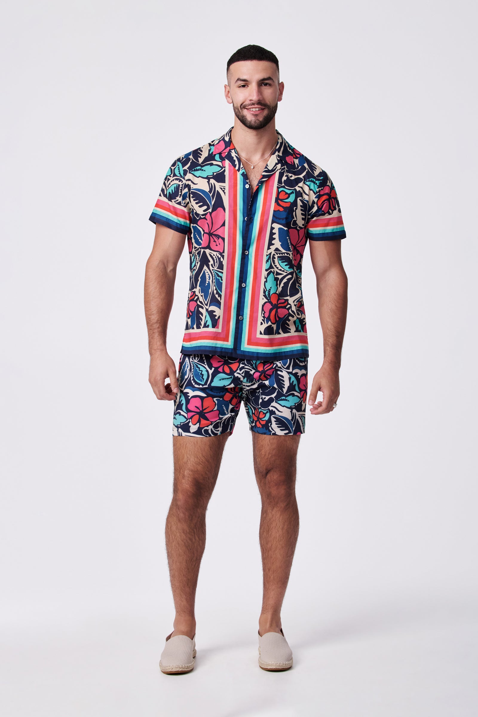 A man smiles, facing forward, wearing the Josue Ss Shirt—a matching short-sleeve button-up with bold, colorful floral and geometric patterns—and beige slip-on shoes, standing against a plain white background.