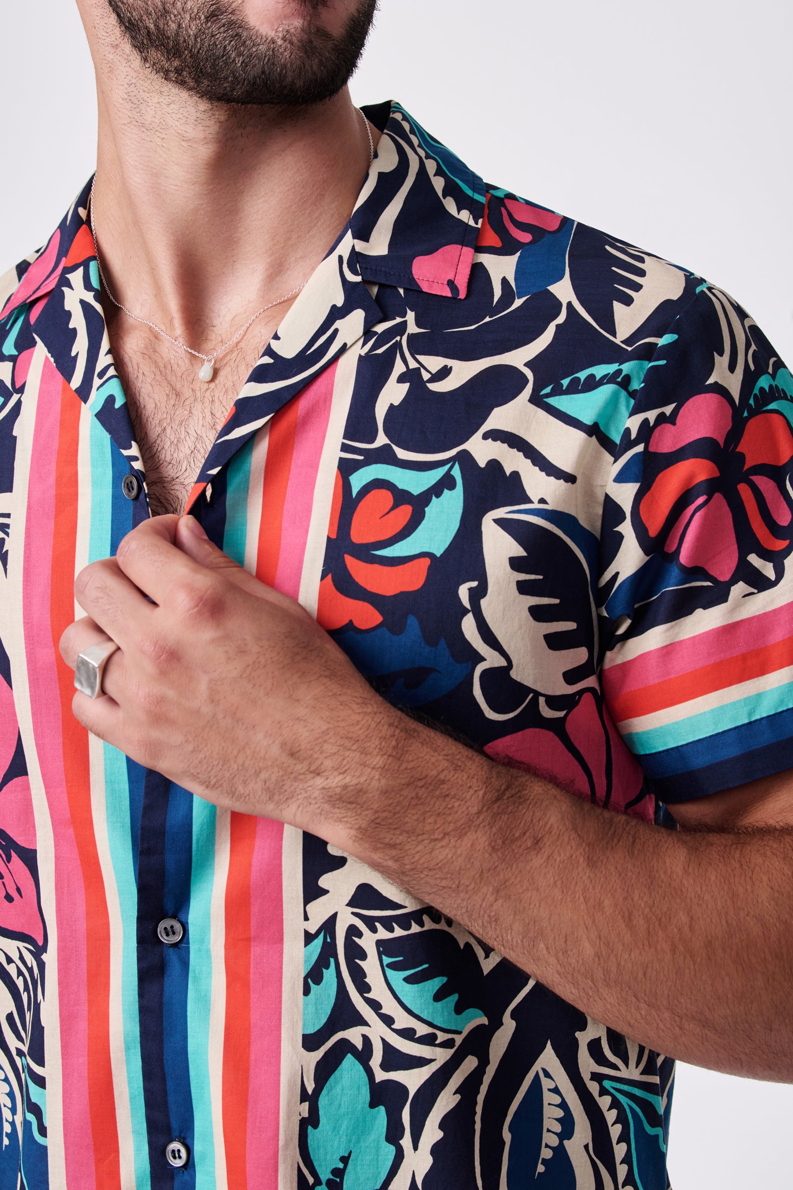 A man wearing the Josue Ss Shirt, a colorful short-sleeved button-up with bold red, blue, pink, teal, and cream floral and striped patterns. He holds the collar, displaying a silver ring and thin necklace.