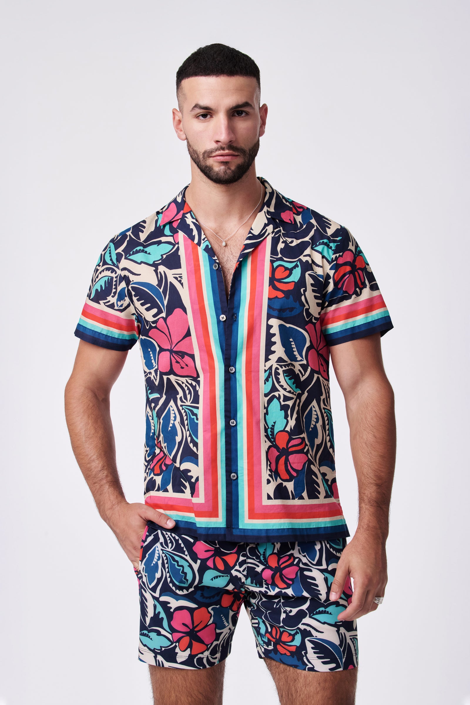 A man with short dark hair and a beard stands against a plain background, wearing the Josue Ss Shirt—a matching short-sleeve button-up with shorts, both featuring a bold, colorful tropical floral pattern and stripes.