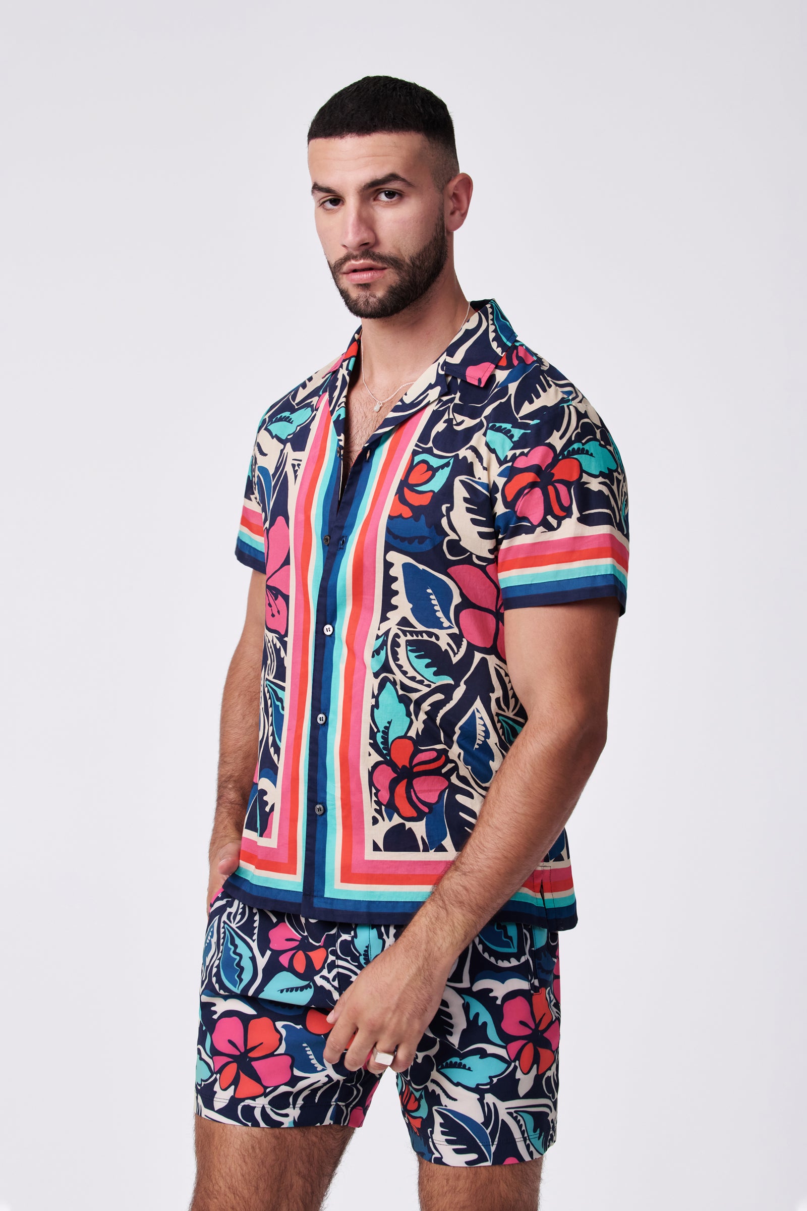 A man with short dark hair and a beard wears the Josue Ss Shirt, a colorful tropical print short-sleeve shirt with pink, blue, and teal floral patterns, standing against a plain light background.