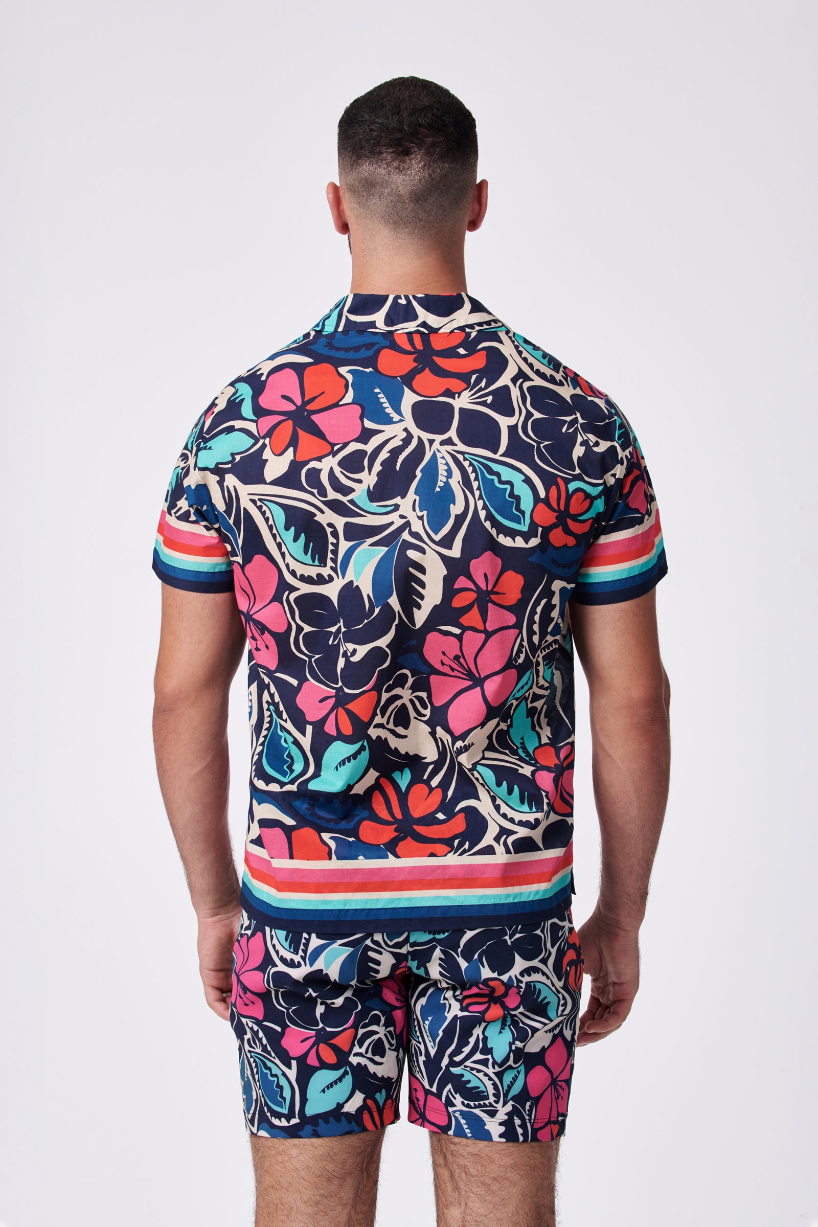 A man stands with his back to the camera in a Josue Ss Shirt and matching shorts, featuring a bold, colorful floral pattern and striped accents on dark fabric. The background is plain white.