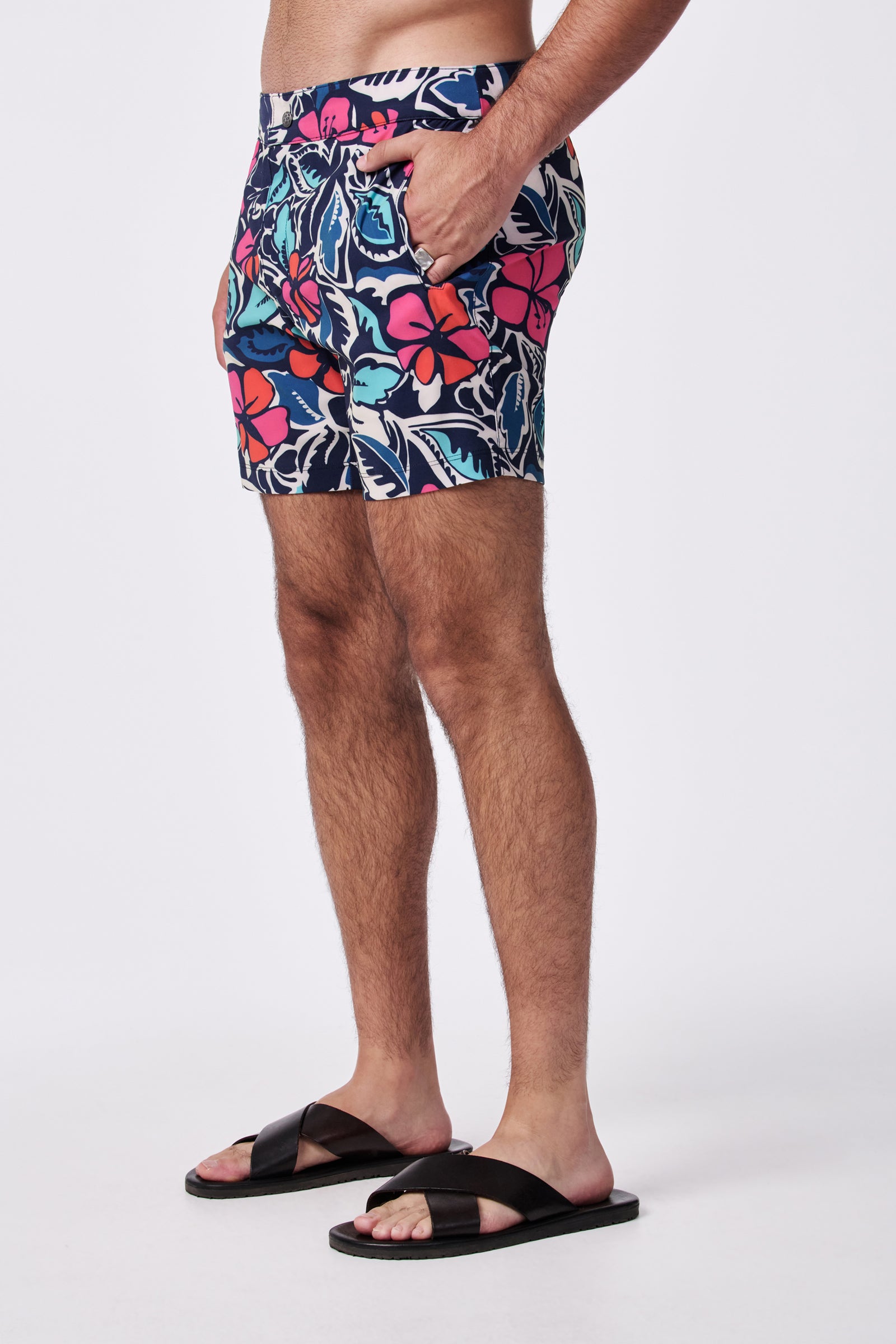 A person stands sideways with hands in pockets, wearing the Honolulu Board Short - Copacabana and black sandals against a plain white background.