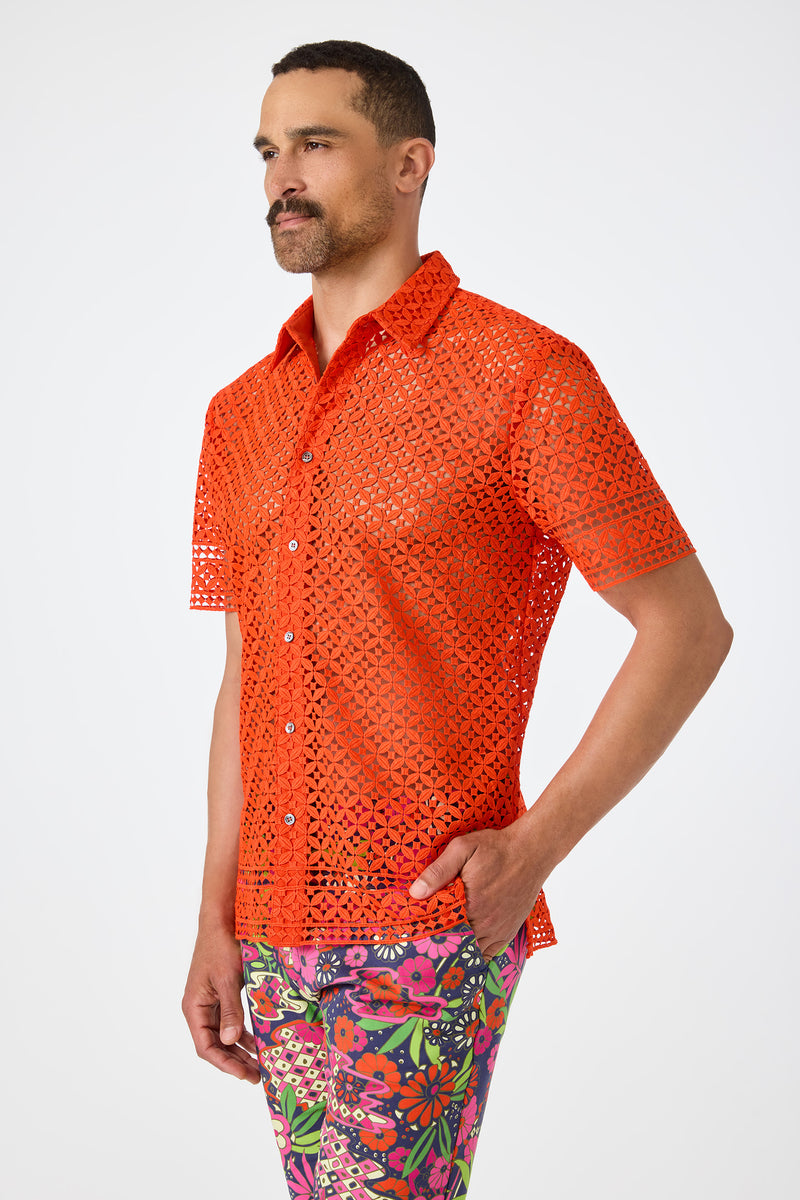 A man with a short beard poses against a white background, wearing the Belmont Shore Shirt‚Äö√Ñ√Æa bright orange, short-sleeved, see-through lace shirt‚Äö√Ñ√Æand colorful floral pants, with one hand in his pocket.