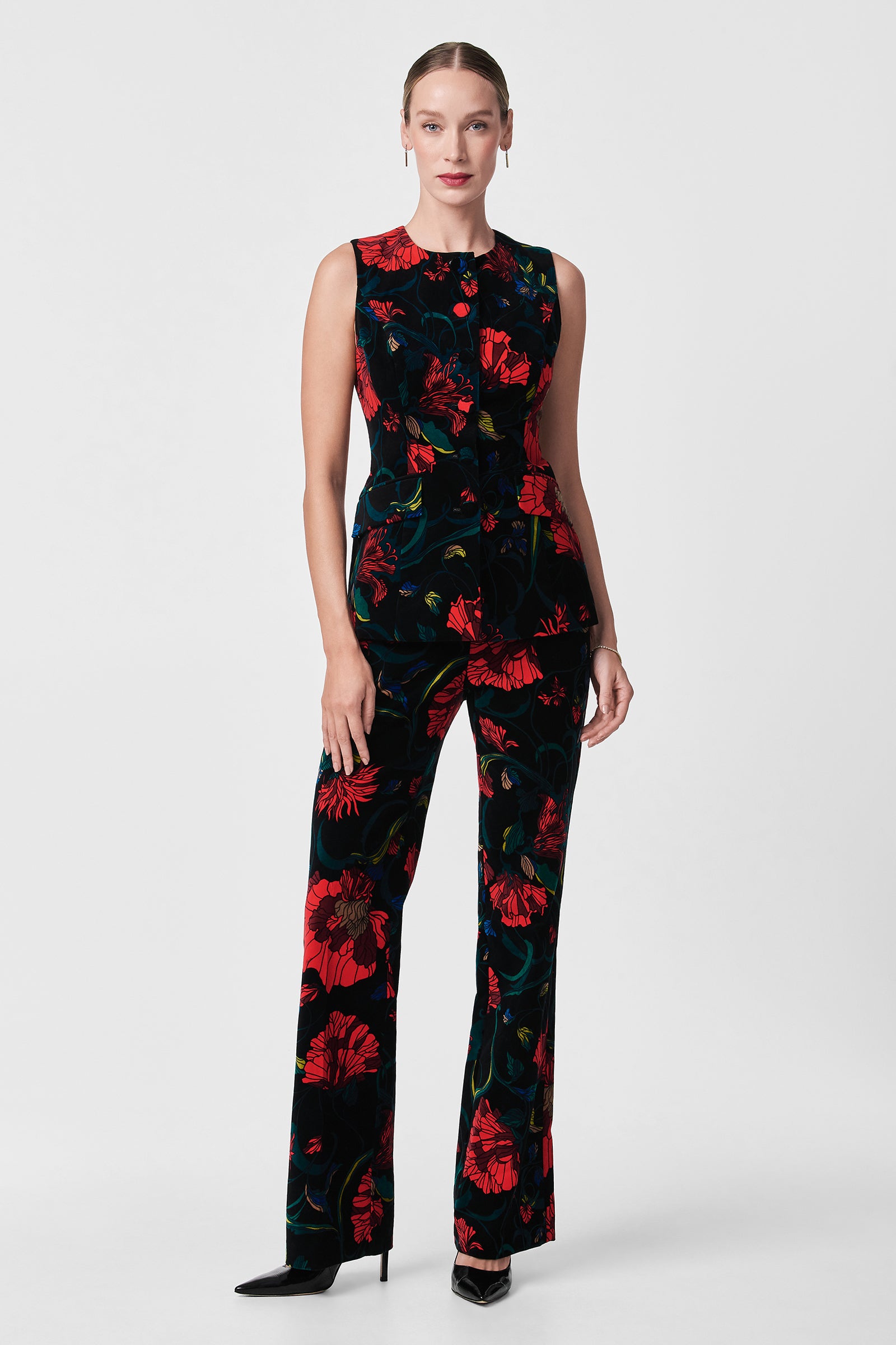A woman faces forward in the Kary Printed Velvet Vest, a sleeveless black garment with a bold red floral pattern, paired with black heels against a plain light gray background.