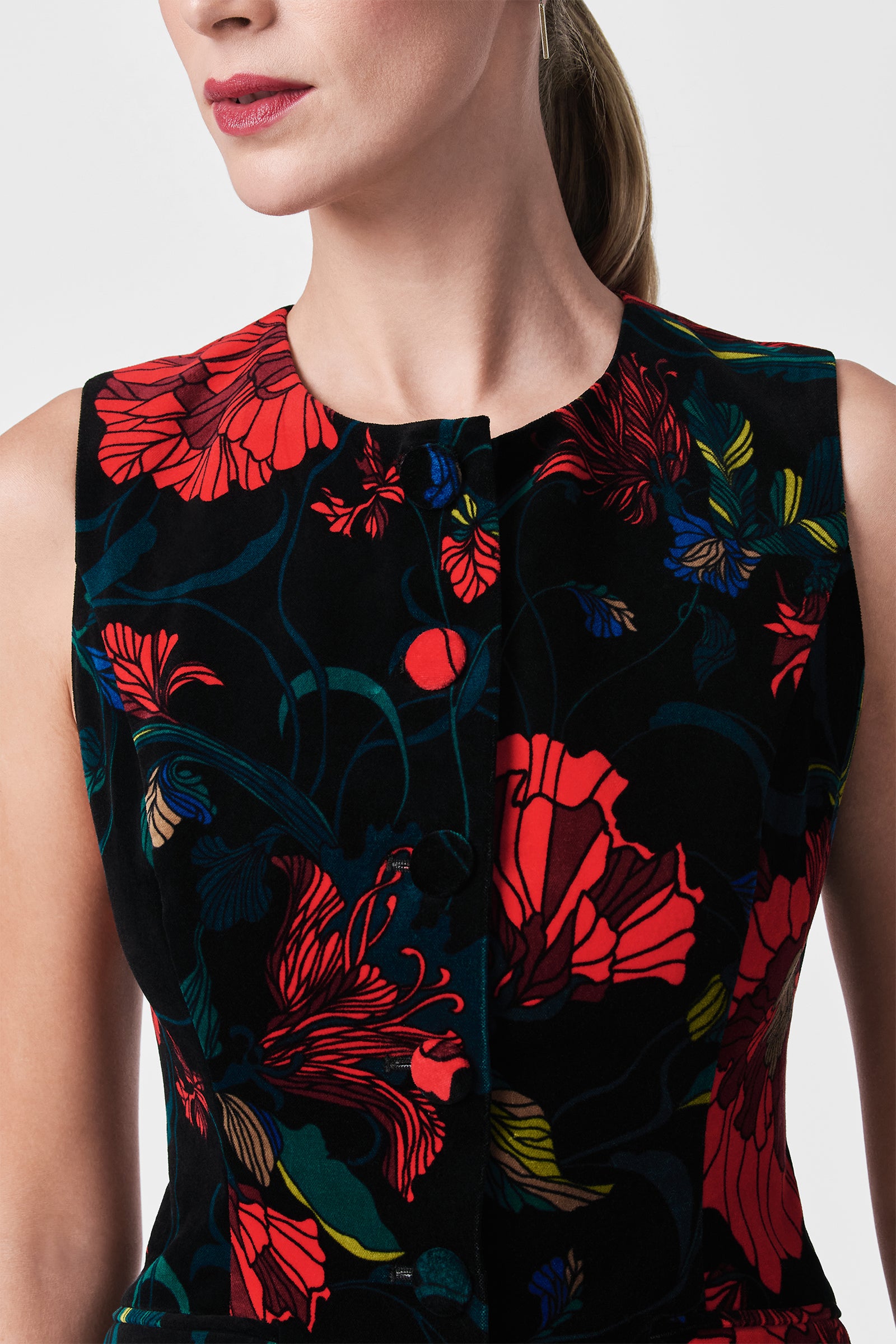 A woman wears the Kary Printed Velvet Vest, a sleeveless black top adorned with bold red, blue, and green floral and leafy patterns. The image highlights her upper torso and the detailed prints on the vest.