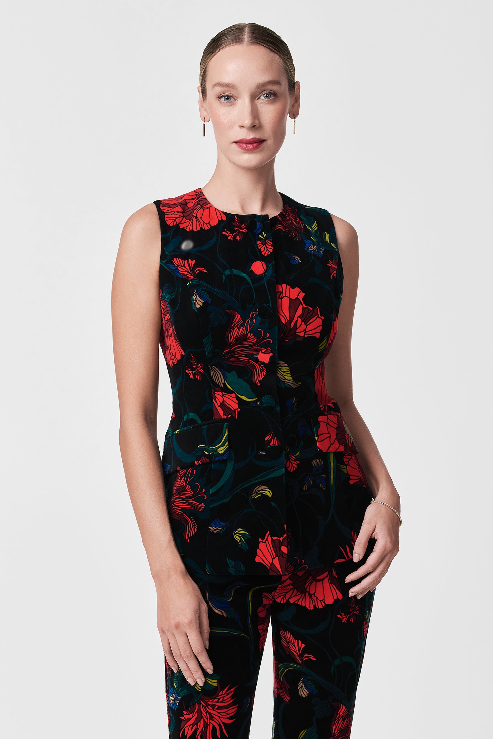 A woman with light skin and blonde hair in a sleek bun models the Kary Printed Velvet Vest, featuring a striking red and green floral pattern, against a plain white background.