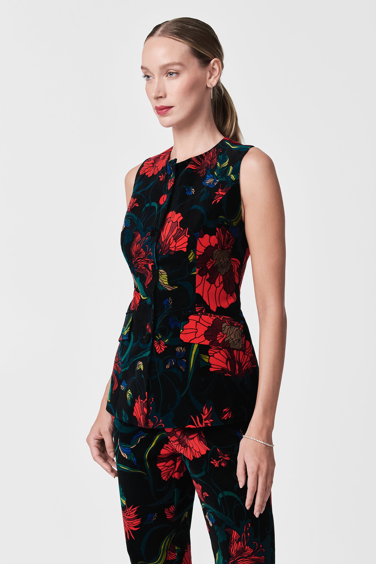 A woman with light skin and straight, pulled-back hair wears the Kary Printed Velvet Vest and matching pants in a bold red and blue floral pattern, standing against a plain white background.