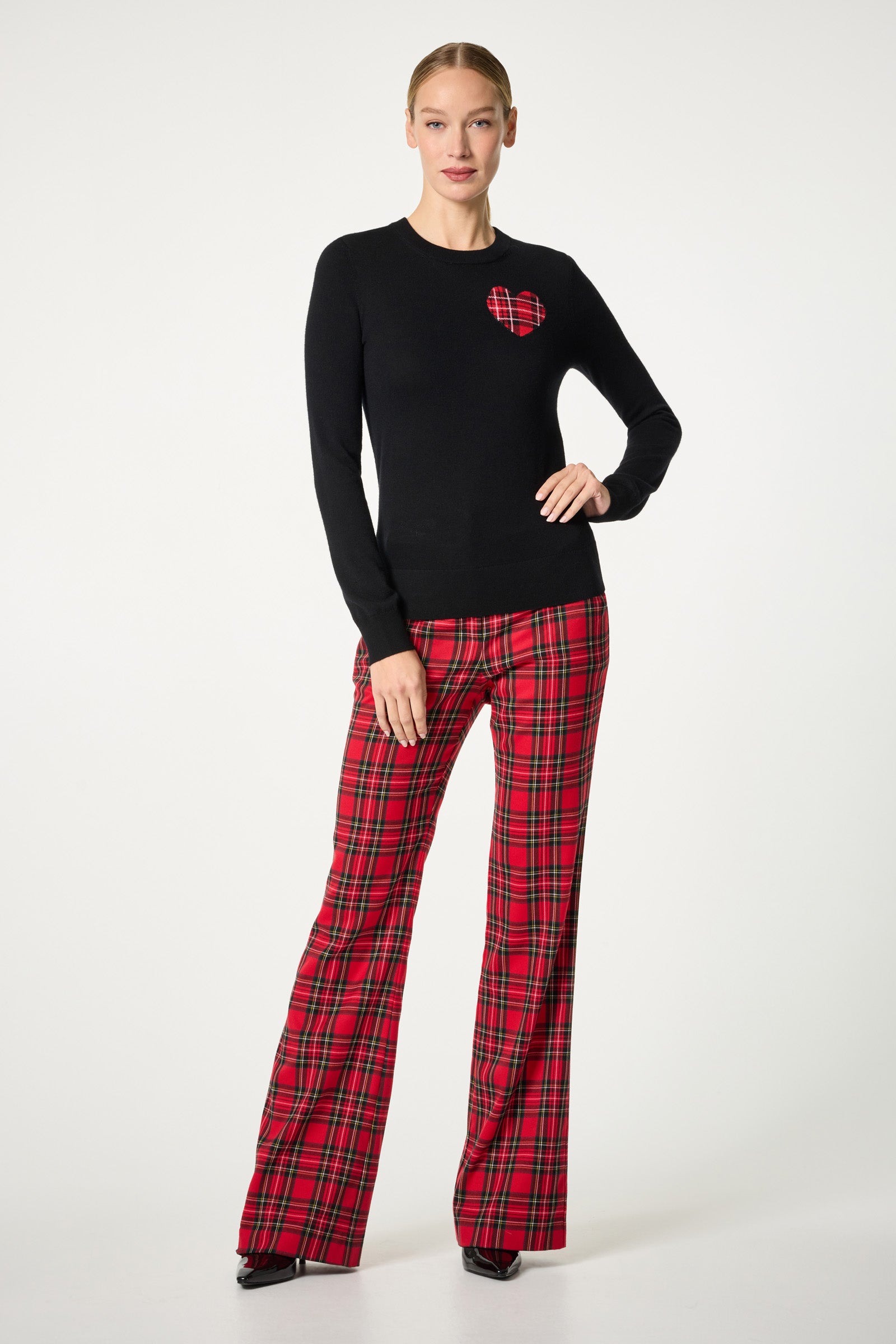 A woman with her hair tied back stands against a white background wearing the Plaid Heart Crewneck Sweater, which features a red plaid heart on the chest, paired with red plaid flared pants and black pointed shoes.