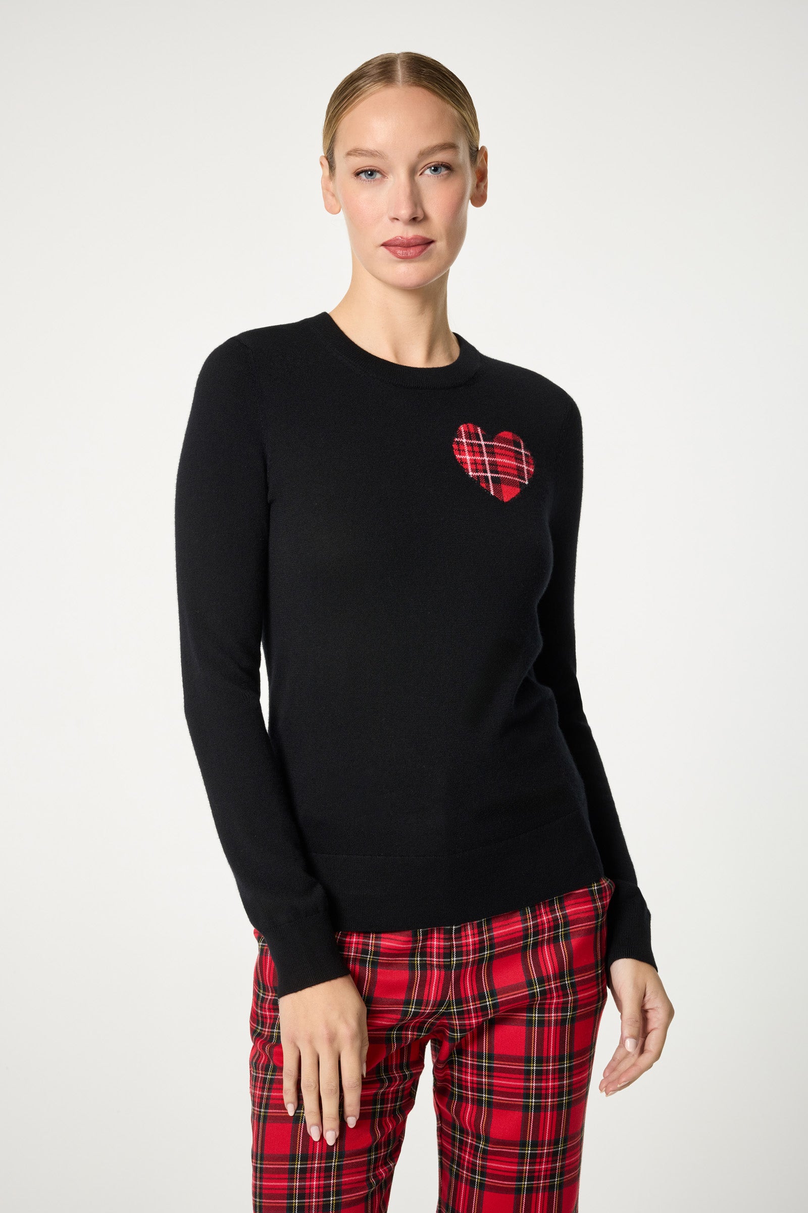 A person wearing the Plaid Heart Crewneck Sweater, featuring a red plaid heart on a black background, and matching red plaid pants, stands against a plain light backdrop.