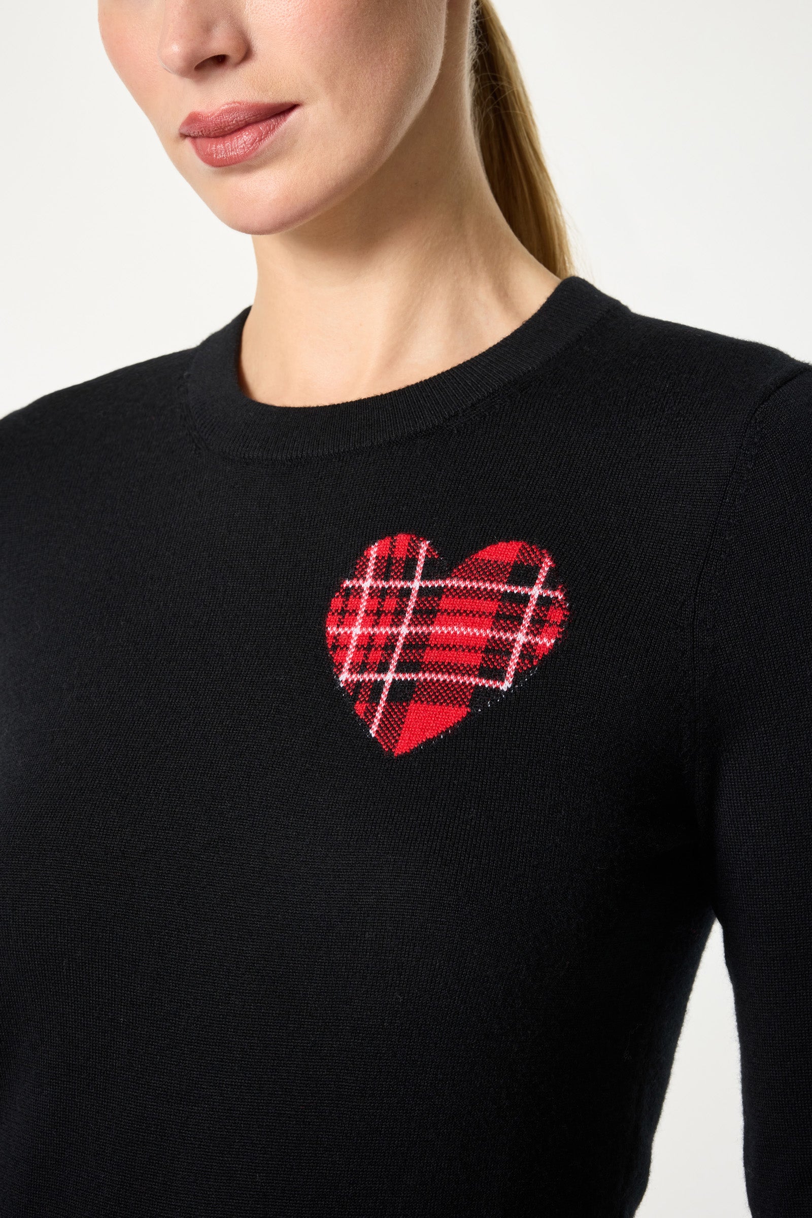 A woman wears the Plaid Heart Crewneck Sweater, a black top with a red and black plaid heart on the upper left chest. Her hair is tied back, and her face is partially visible.
