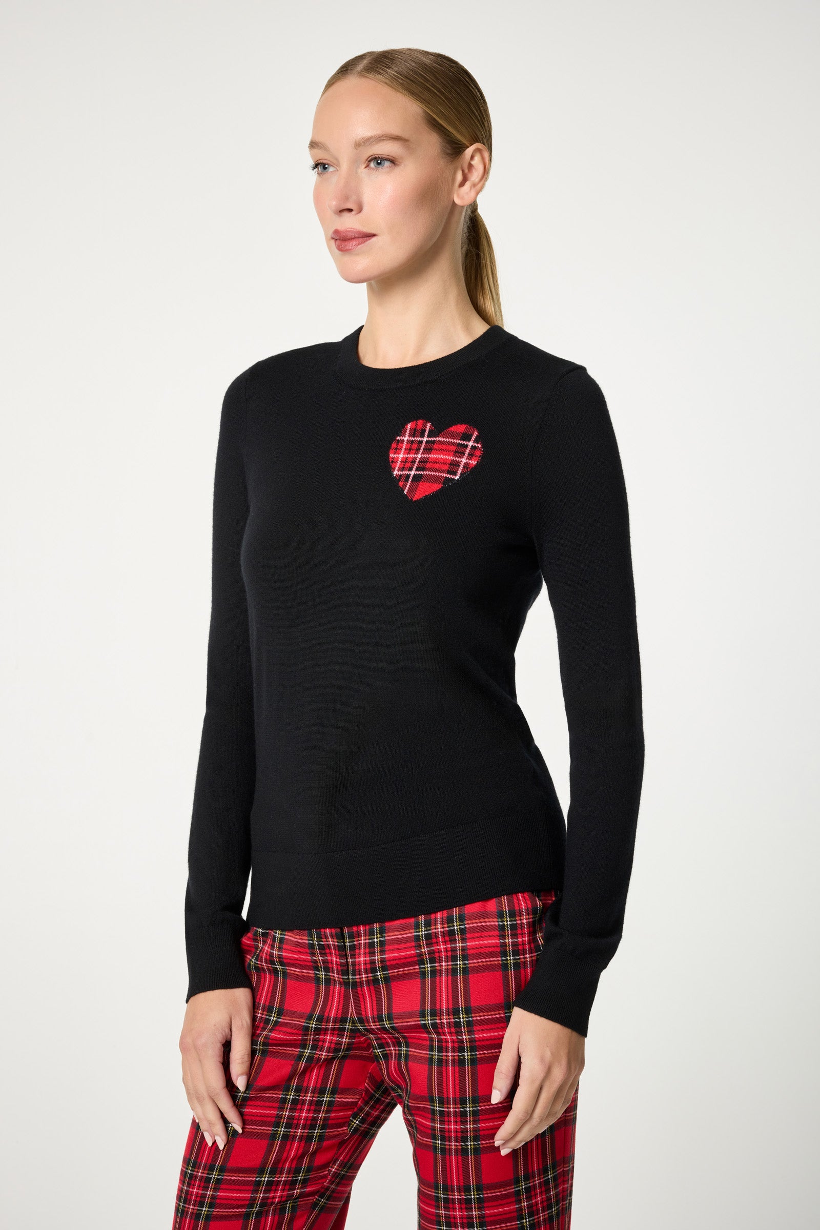 A woman wears the Plaid Heart Crewneck Sweater, a black top with a red plaid heart patch on the chest, paired with red plaid pants, standing against a plain white background.