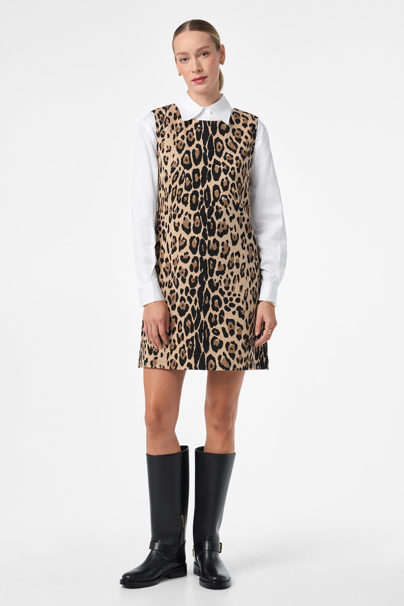 A woman stands facing forward in the Adapted Dress—a sleeveless, knee-length leopard print style over a white collared shirt. She wears black knee-high boots, with her hair tied back, set against a plain white background.