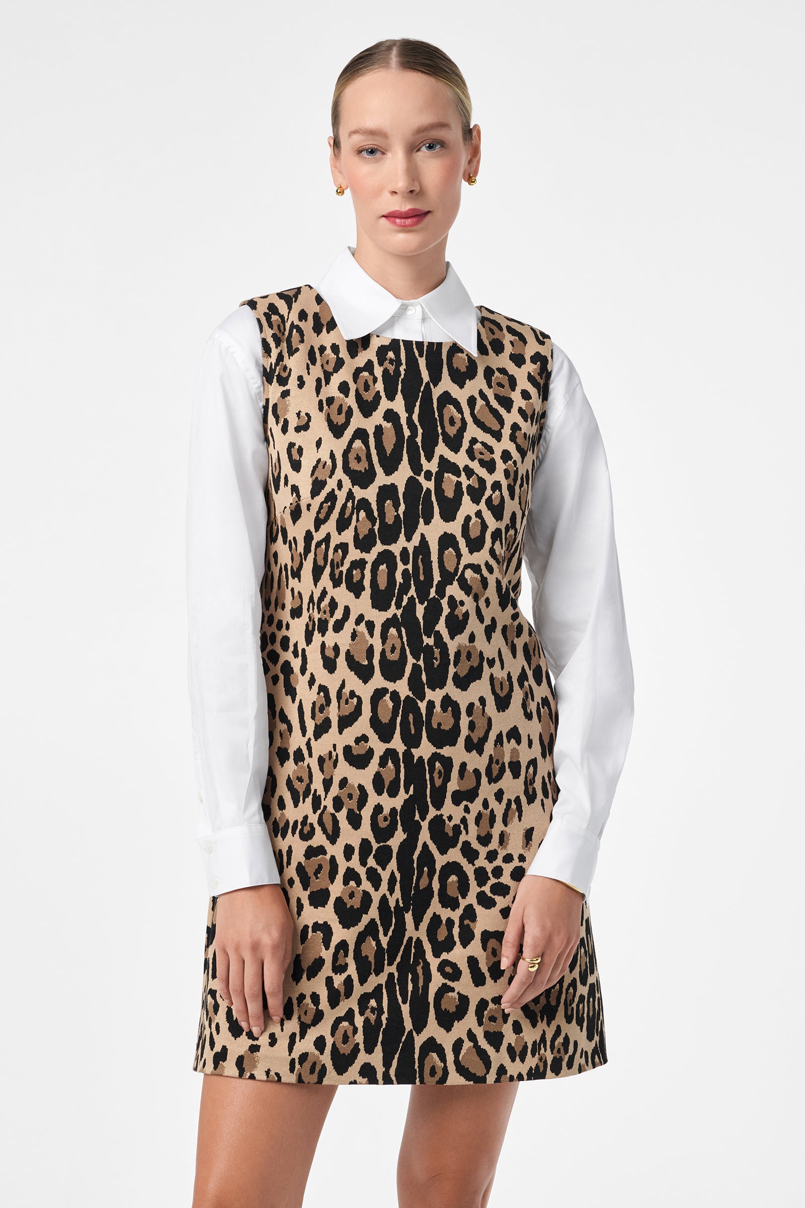 A woman in the Adapted Dress—a sleeveless, leopard-print style over a white collared shirt—stands against a white background. She has blonde hair pulled back and smiles slightly at the camera.