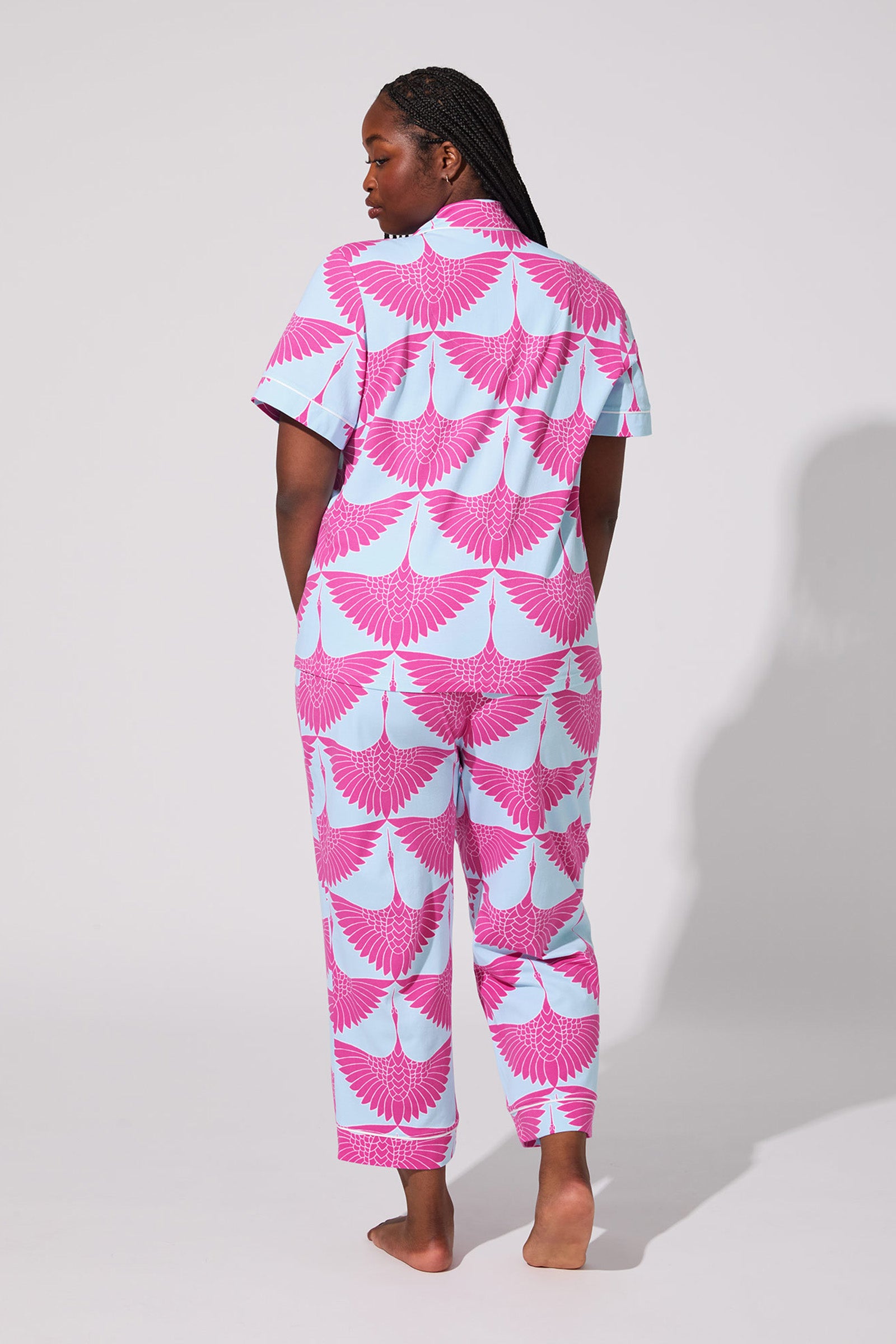 A person stands barefoot facing away in the Cranes Crop PJ Set—light blue, short-sleeve and pant pajamas with a bold pink and white crane bird pattern—against a plain, light background.