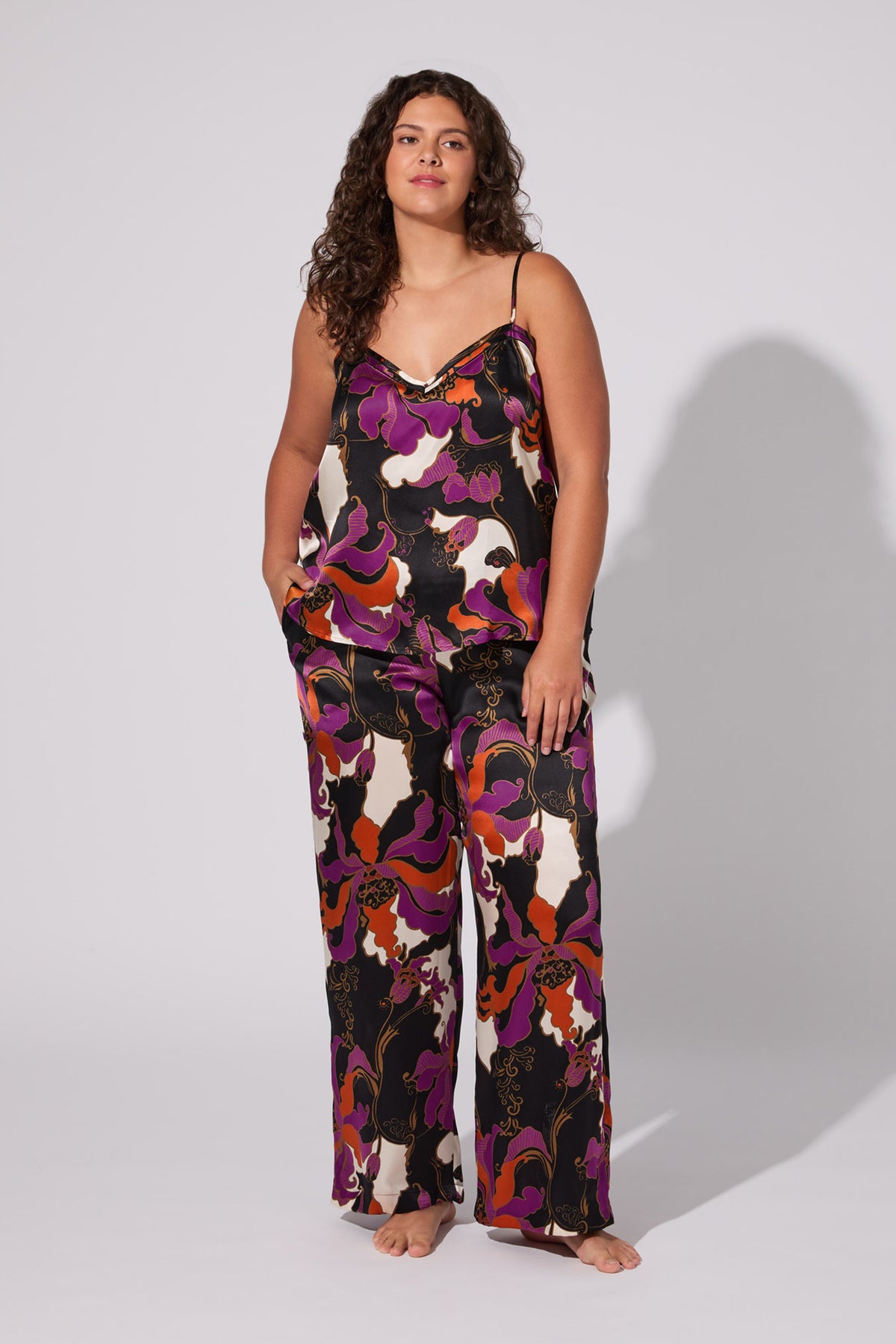 A woman with long curly hair models the Fall Floral Silk Cami Pj Set—colorful, loose-fitting camisole and matching wide-leg pants with a bold floral pattern—standing barefoot against a plain, light background.