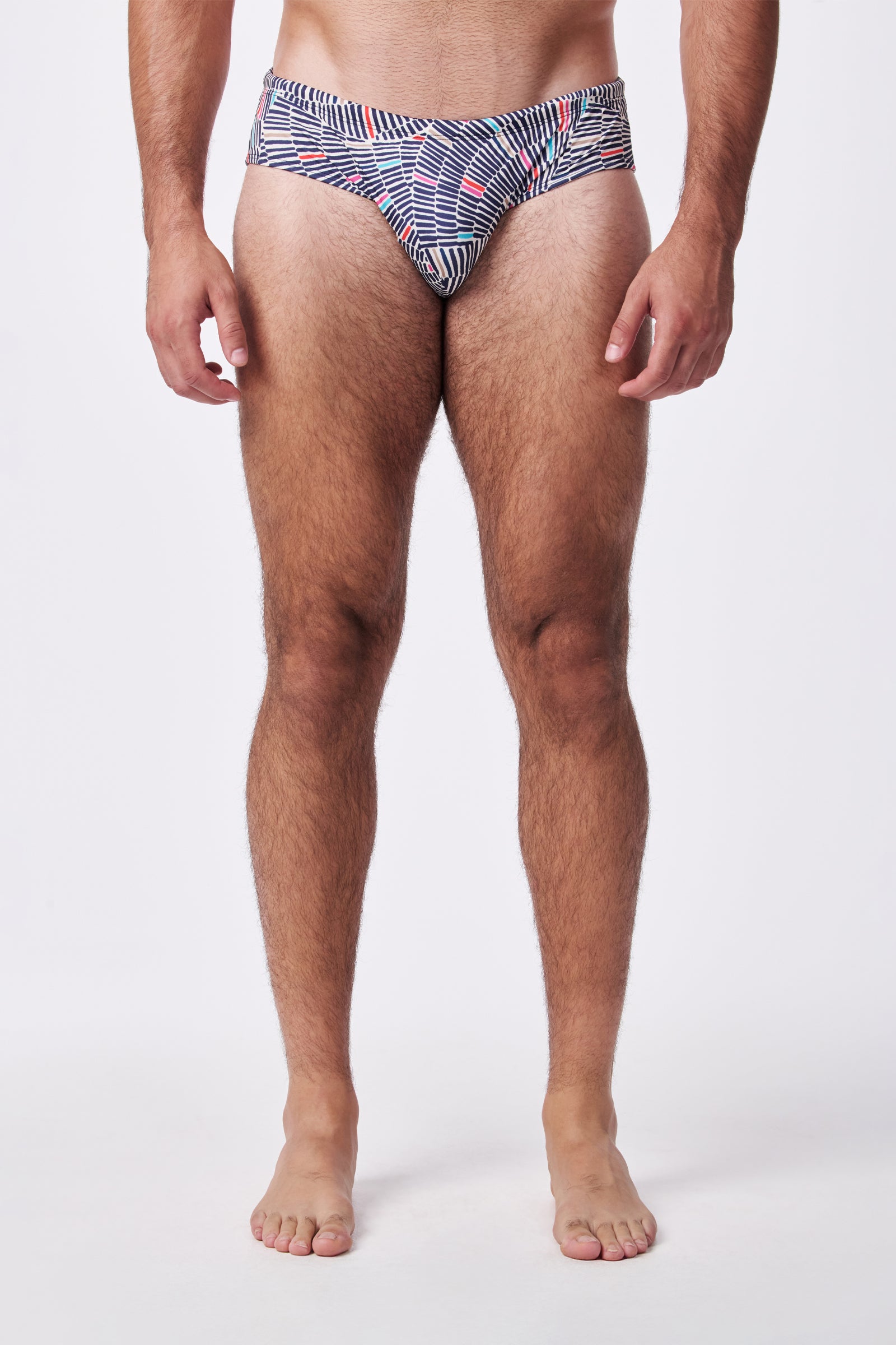 A man stands barefoot against a plain white background, wearing the Bondi Swim Trunk featuring a blue, white, and red geometric pattern. His arms are at his sides, and his face is not shown.