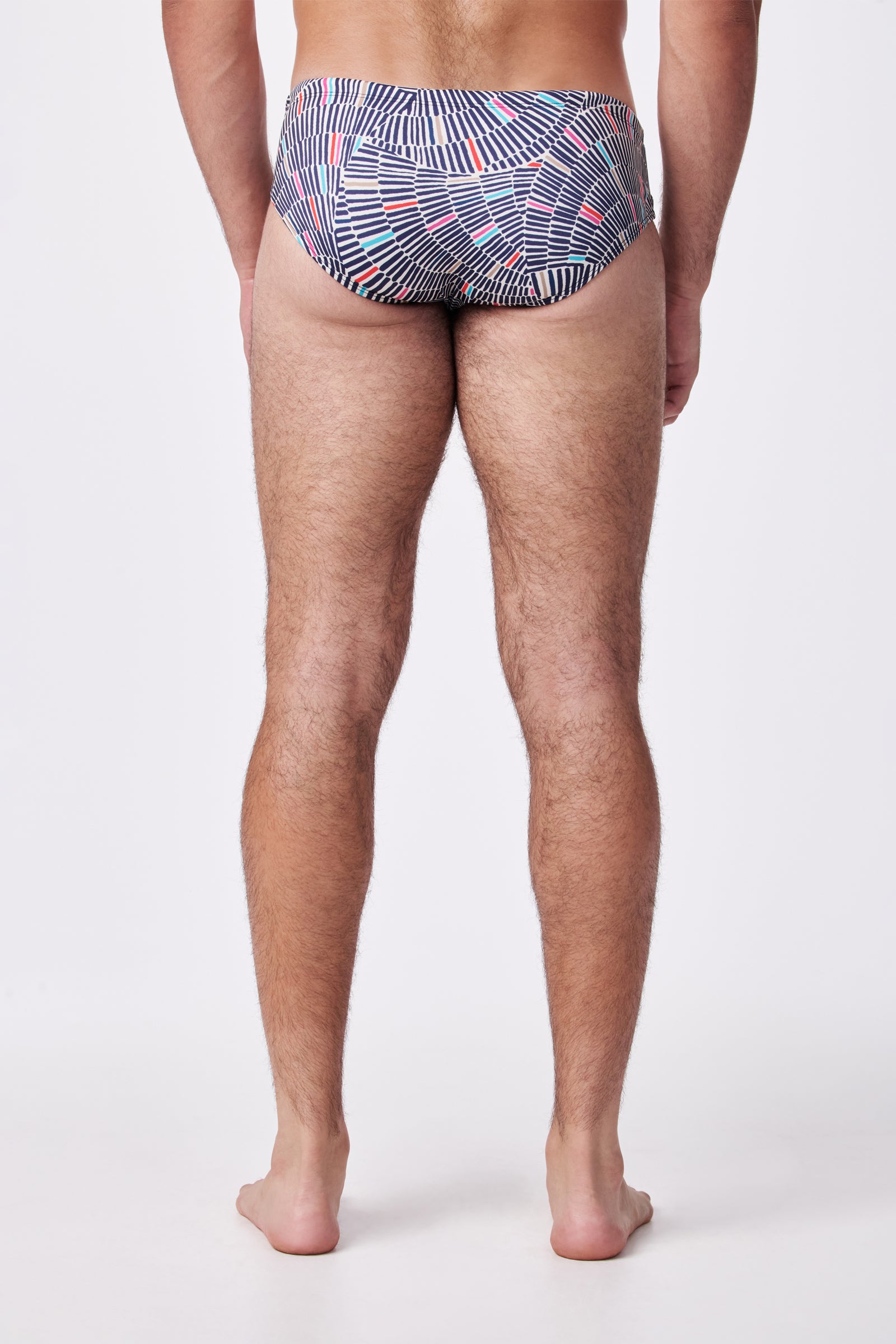 A man stands barefoot with his back to the camera, wearing the Bondi Swim Trunk featuring a blue, white, and red geometric pattern. The background is plain white.