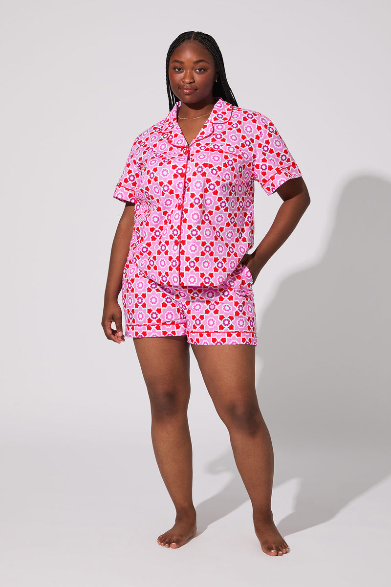 A woman stands barefoot against a plain background, wearing the Mosiac Tile Short Pj Set—a matching pink and red patterned short-sleeve pajama set with shorts. She has one hand in her pocket and looks confidently at the camera.