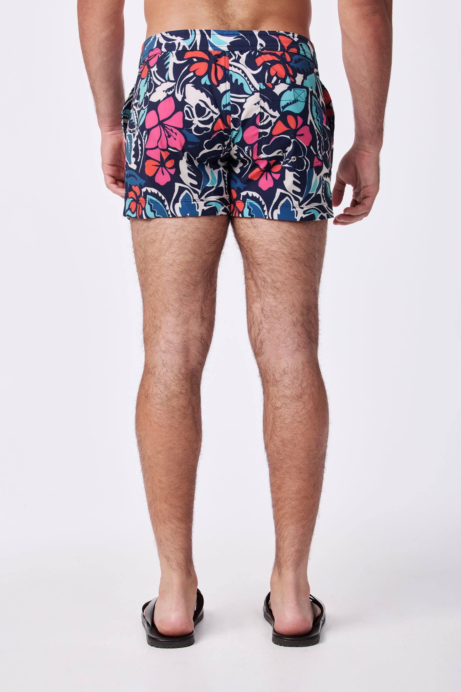 A person wearing the Chico Swim Trunk - Copacabana, featuring a colorful floral pattern, and black flip-flops stands with their back to the camera against a plain white background.