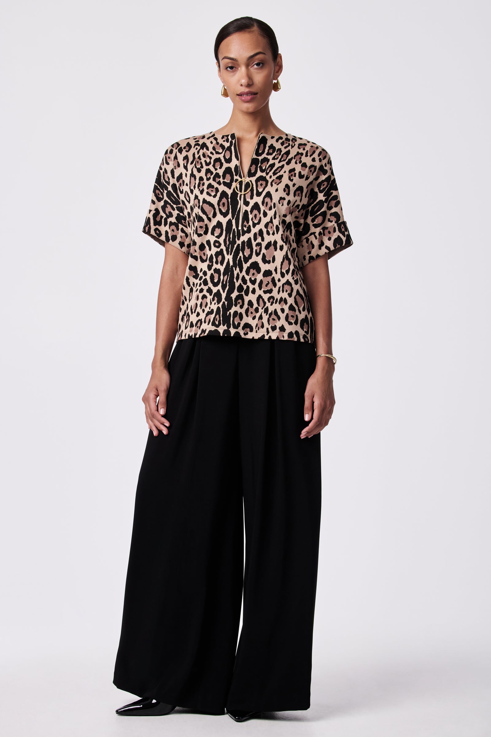 A woman stands against a plain background, wearing the Stagehands Half-Zip Top— a short-sleeve leopard print blouse with a front zipper—paired with wide-leg black pants. She has her hair pulled back and wears earrings and a bracelet.