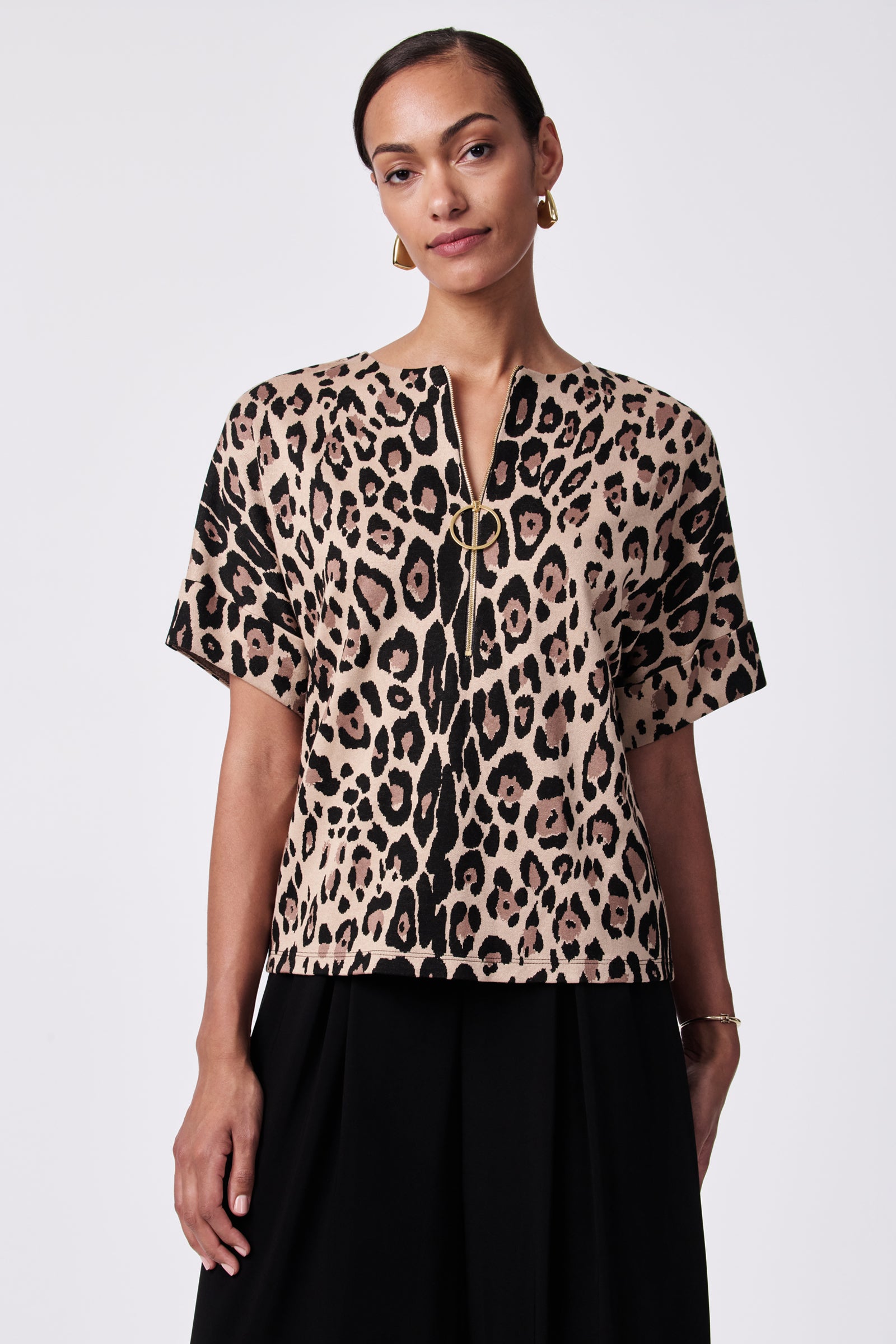 A woman wears the Stagehands Half-Zip Top, a short-sleeved beige and black leopard print top with a front zipper, paired with a black skirt. She accessorizes with gold hoop earrings and a minimalist bracelet against a white background.