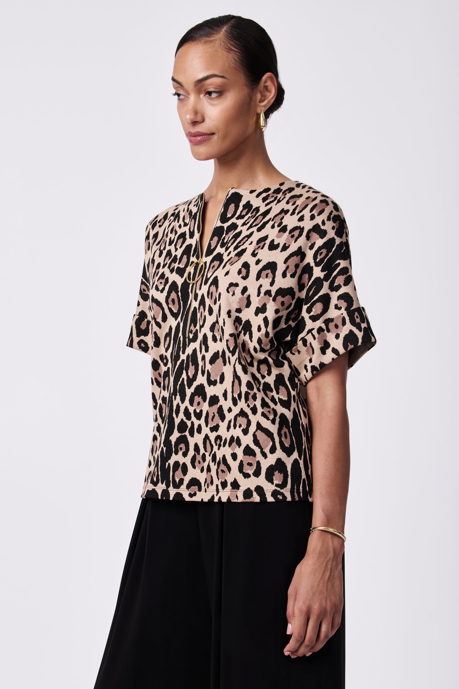A woman stands against a plain background wearing the Stagehands Half-Zip Top, featuring a beige and black leopard print, short sleeves, paired with black pants and gold accessories, looking slightly to the side with a soft smile.