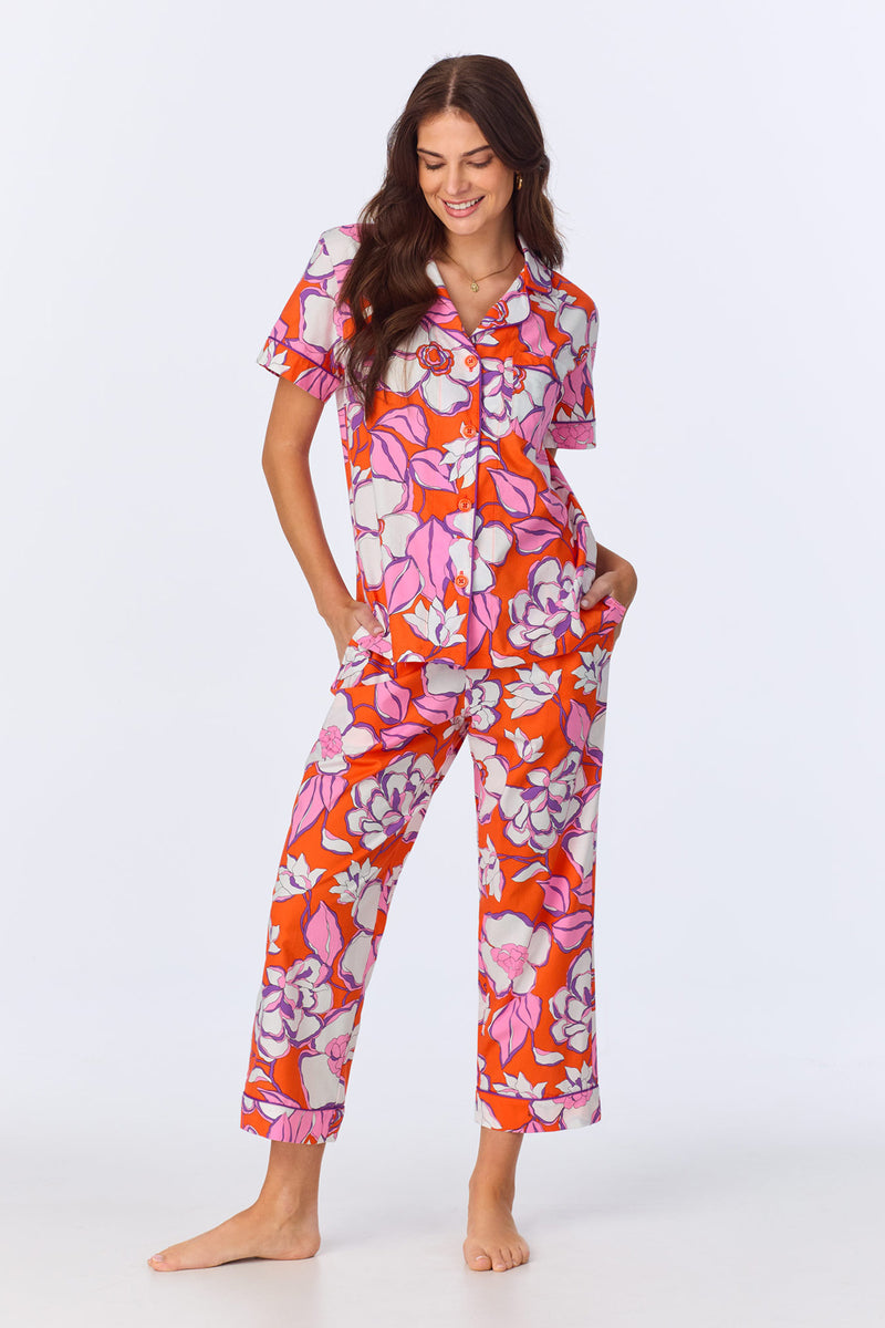 A woman stands barefoot in the Crop Classic PJ Set - Gardenia Poplin, a multi-colored floral pajama with short sleeves and long pants. She smiles with her hands in her pockets against a plain white background.