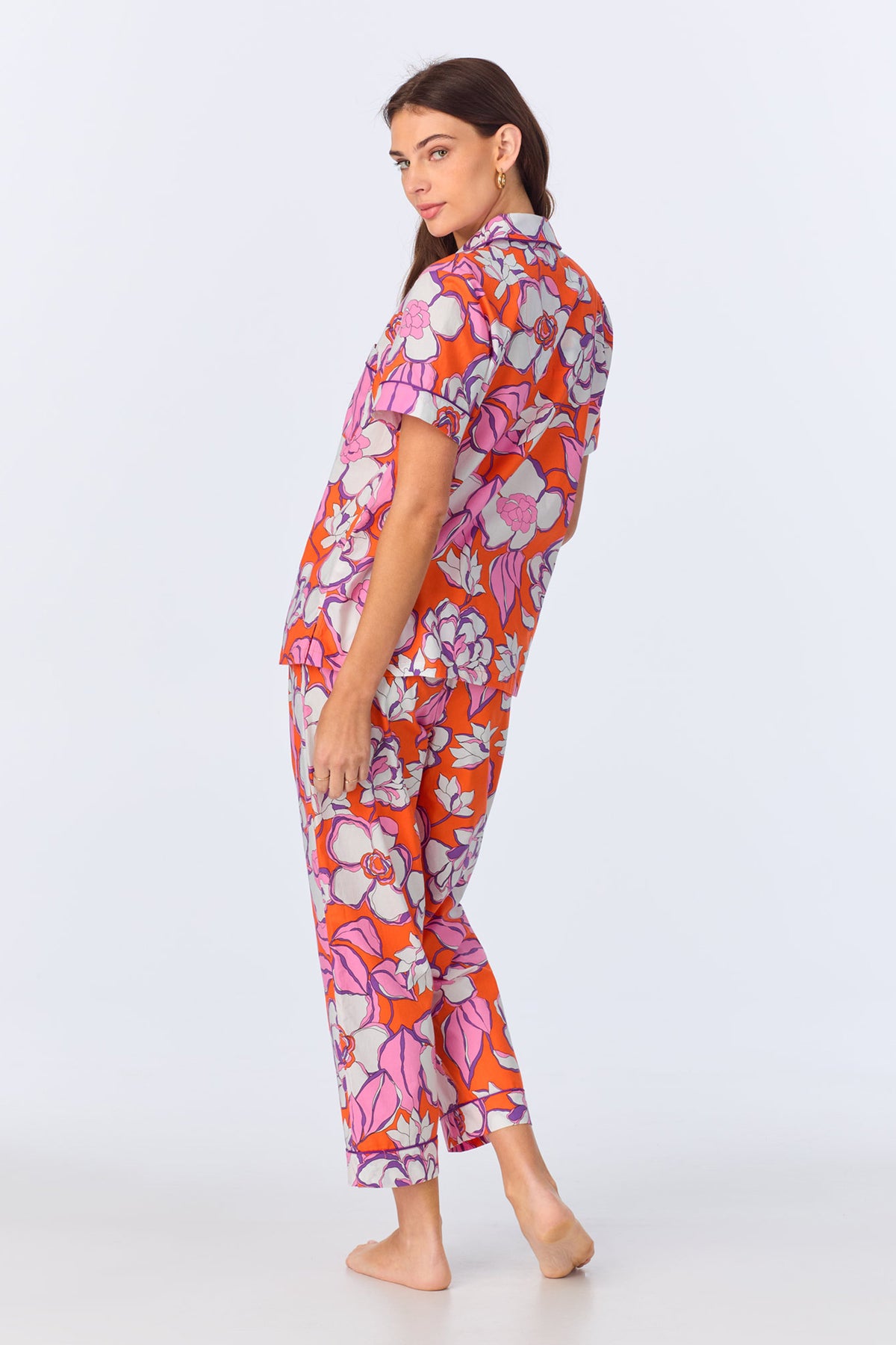 A woman stands barefoot, turned slightly to the side, wearing the Crop Classic PJ Set - Gardenia Poplin, a vibrant orange pajama set with bold pink, purple, and white florals. The set features short sleeves and cropped pants. The background is plain white.