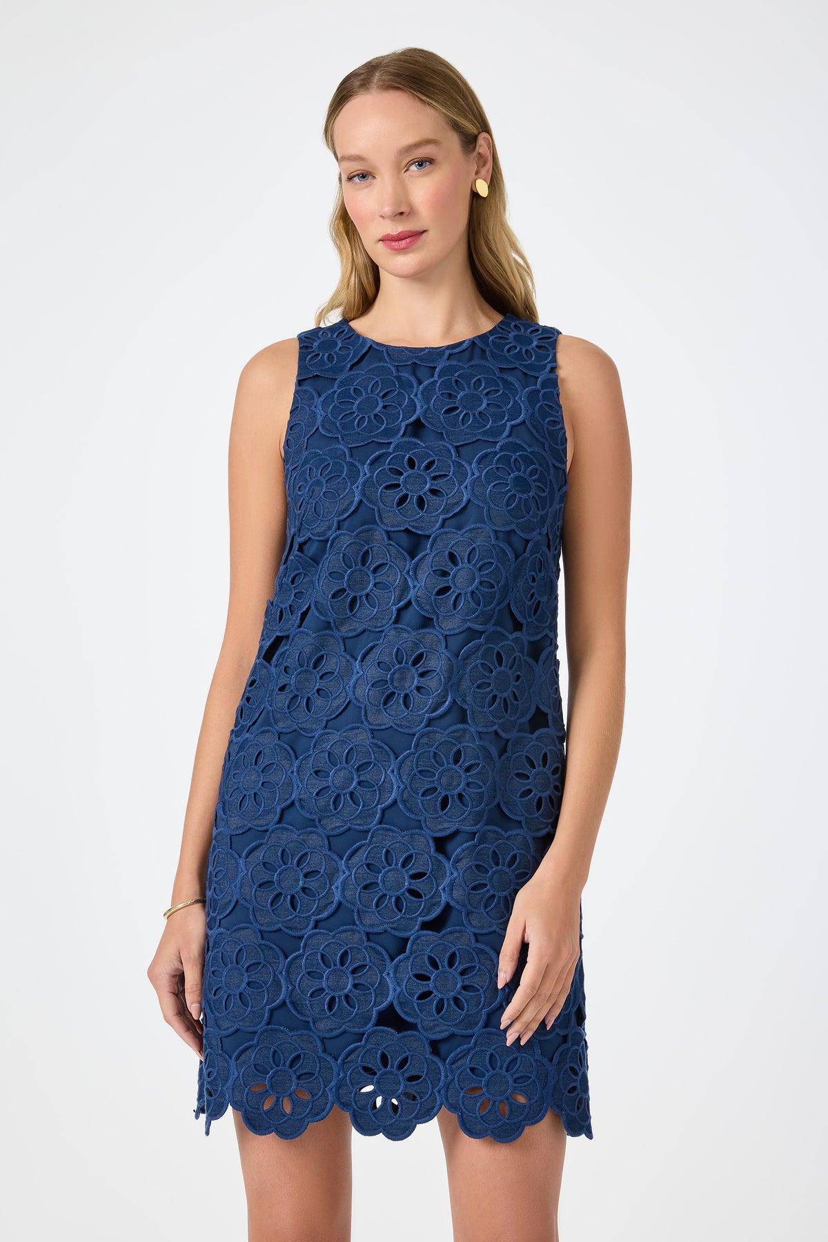 A woman wears the Yakitori Mini Dress - Kusho Embroidery, a sleeveless, knee-length blue dress with bold floral lace patterns, standing against a plain white background. She has long light brown hair and simple accessories.