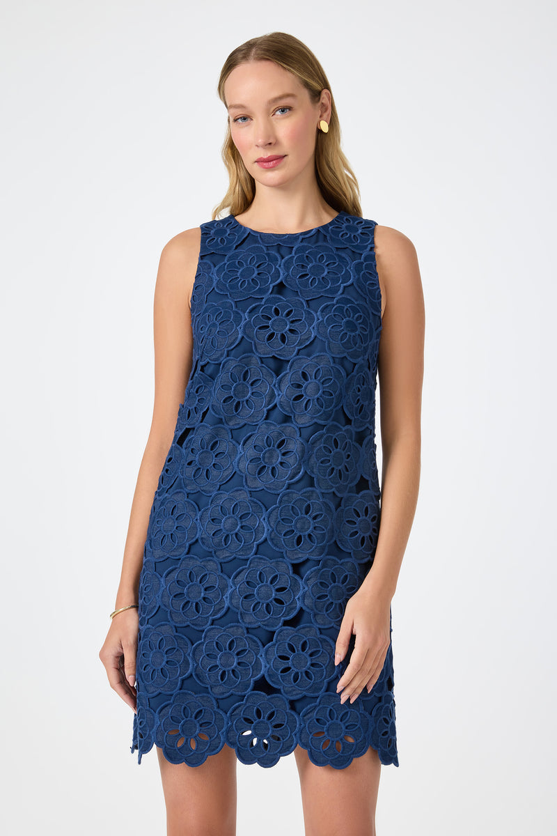A woman wears the Yakitori Mini Dress - Kusho Embroidery, a sleeveless, knee-length blue dress with bold floral lace patterns, standing against a plain white background. She has long light brown hair and simple accessories.