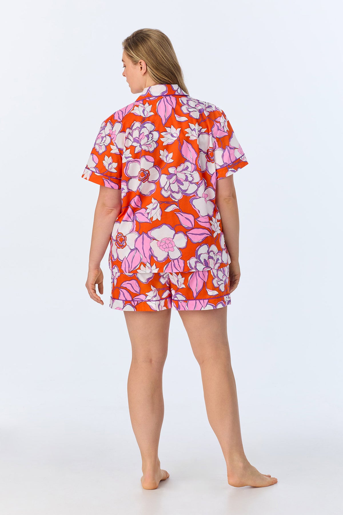A person stands barefoot with their back to the camera, wearing the Shorty PJ Set - Gardenia Poplin, featuring a matching short-sleeved shirt and shorts in a vibrant red, pink, and white floral pattern against a plain light background.
