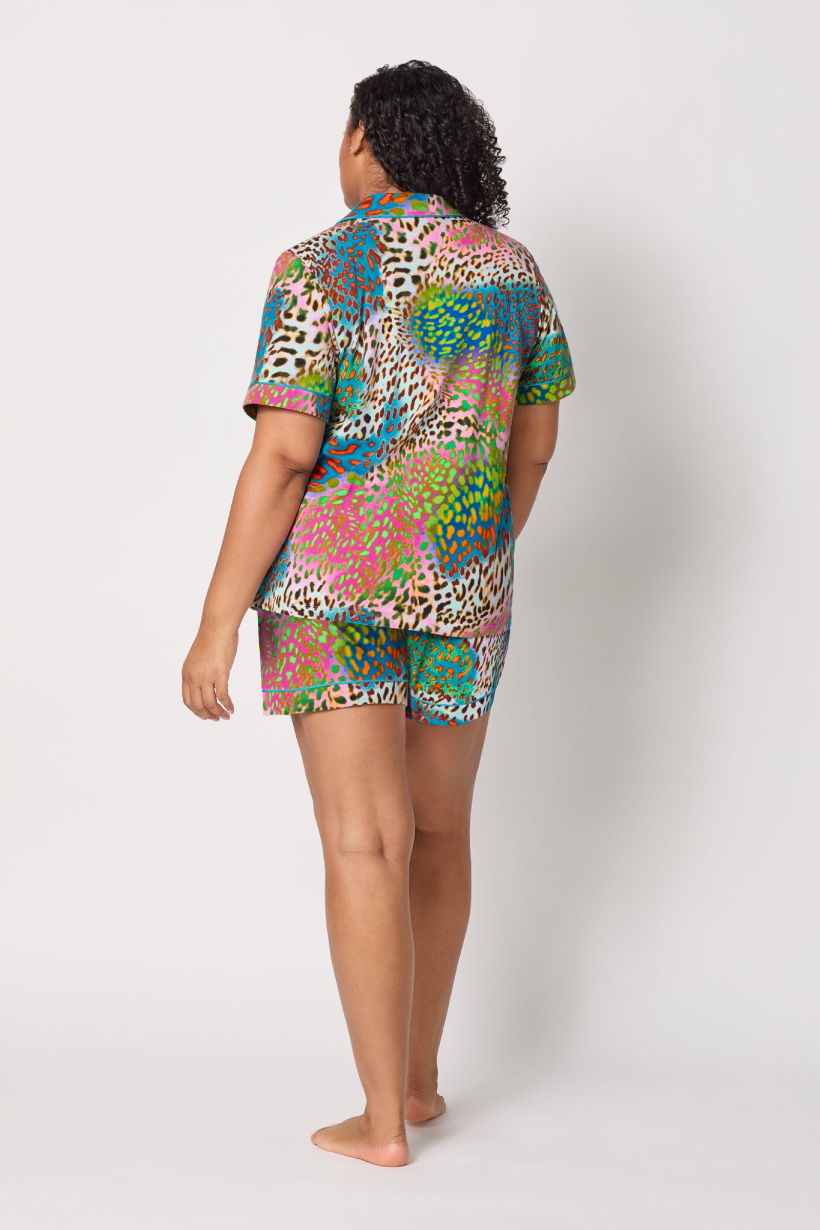 A person stands barefoot facing away, wearing the Shorty PJ Set - Spray Leopard, a colorful, short-sleeved pajama set with a vibrant multicolored animal print. The background is plain white.