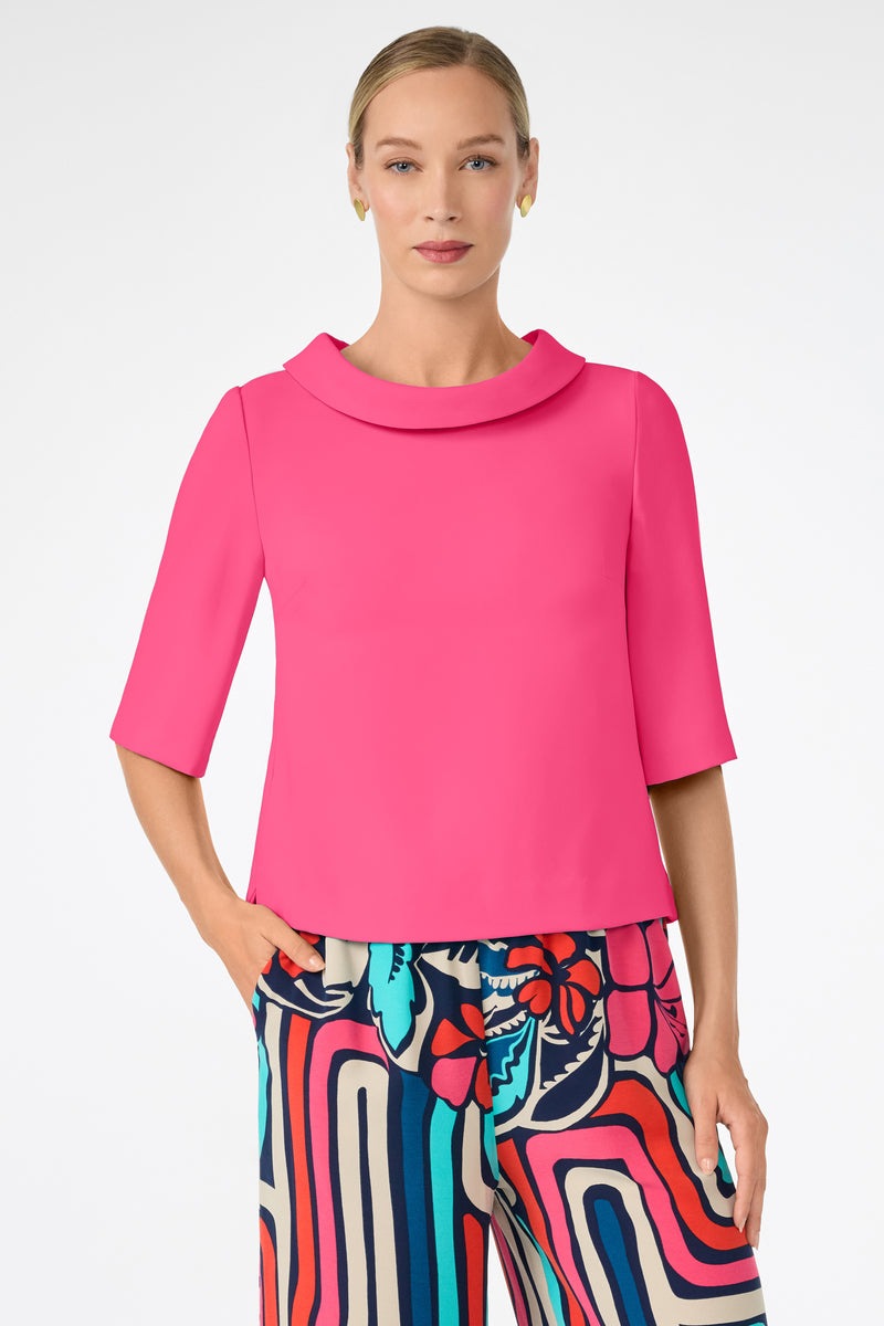 A woman wearing the Kailee Top in bright pink with elbow-length sleeves and a wide collar, paired with colorful abstract-patterned pants in blue, red, and cream, stands against a plain white background.