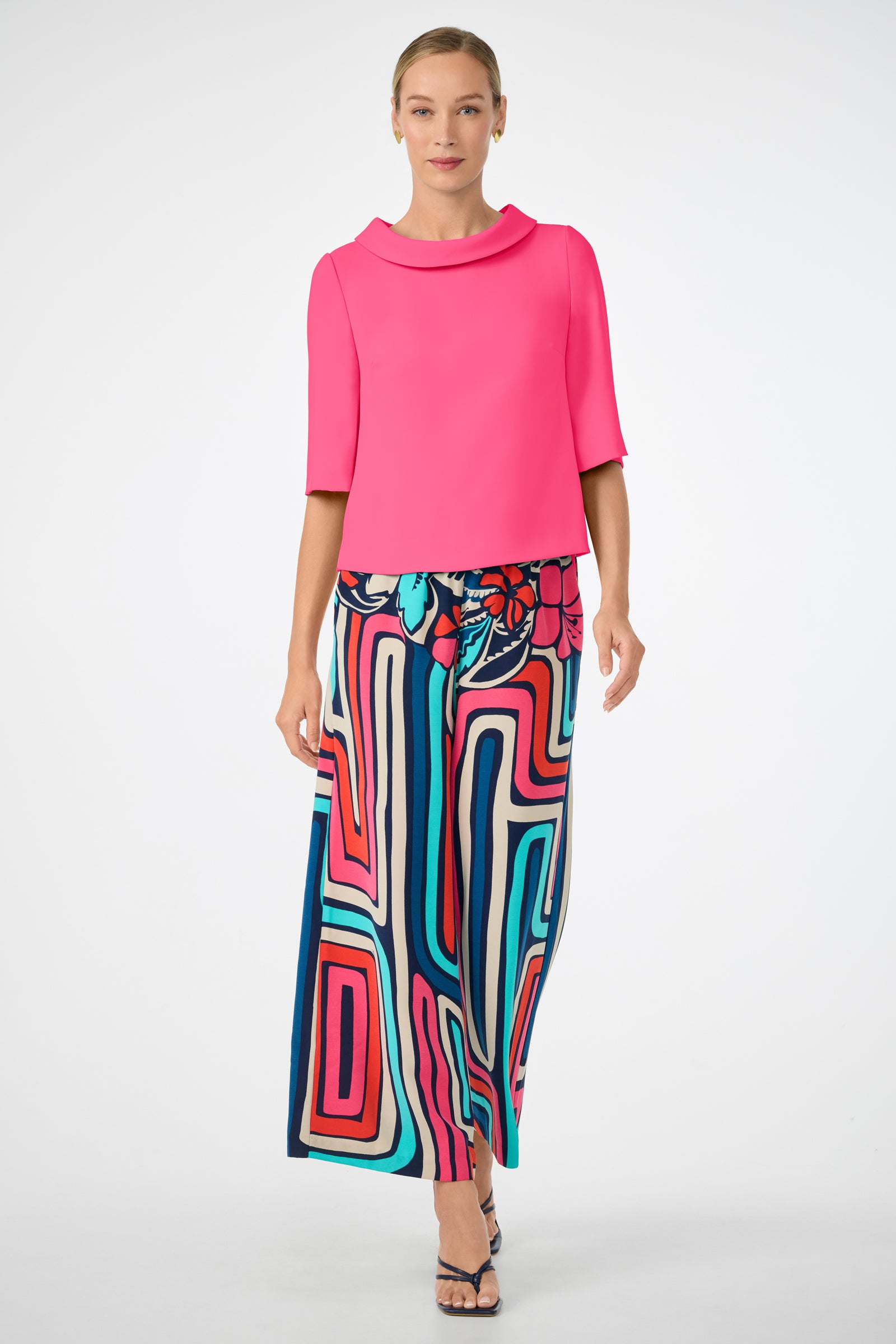 A woman wears the Kailee Top, a bright pink top with elbow-length sleeves, paired with colorful wide-leg pants featuring bold abstract patterns in blue, red, pink, and white. She stands against a plain white background.