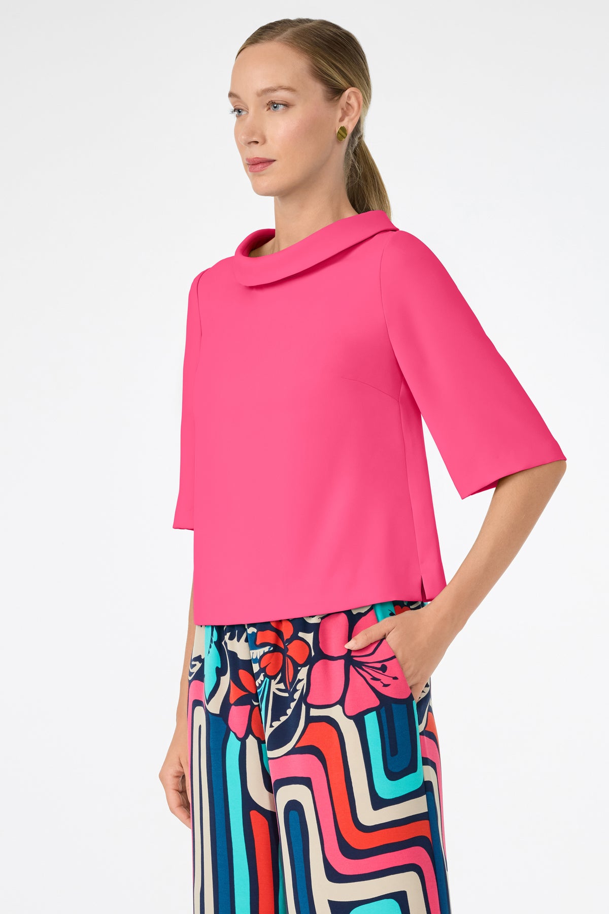 A woman with light skin and straight, blonde hair in a low ponytail wears the bright pink Kailee Top with colorful patterned pants, standing with one hand in her pocket against a plain white background.