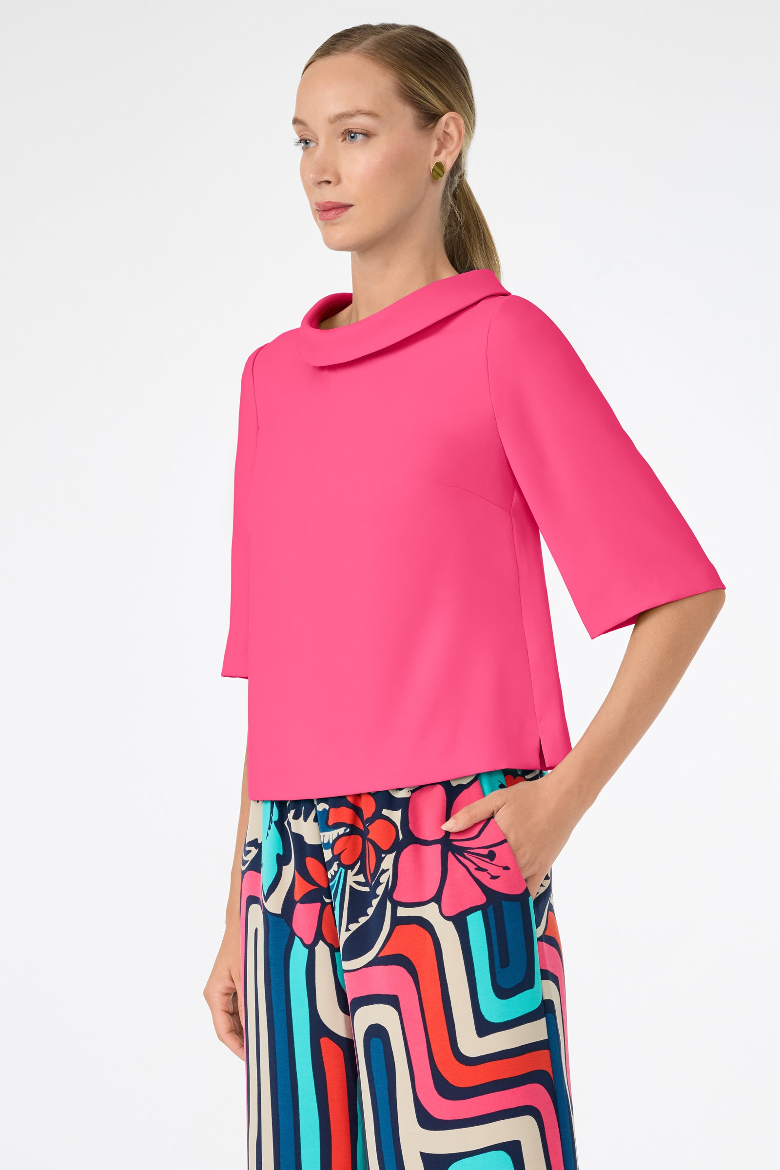 A woman with light skin and straight, blonde hair in a low ponytail wears the bright pink Kailee Top with colorful patterned pants, standing with one hand in her pocket against a plain white background.