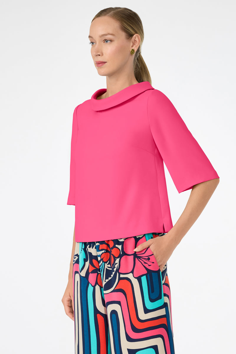 A woman with light skin and straight, blonde hair in a low ponytail wears the bright pink Kailee Top with colorful patterned pants, standing with one hand in her pocket against a plain white background.