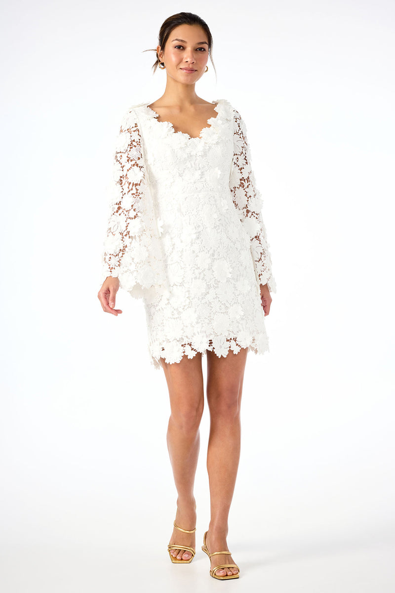 A woman wearing the Ariah Long-Sleeve Dress - Love All Lace in Whitewash, featuring sheer floral sleeves and scalloped edges, paired with gold strappy heels, stands against a plain white background.