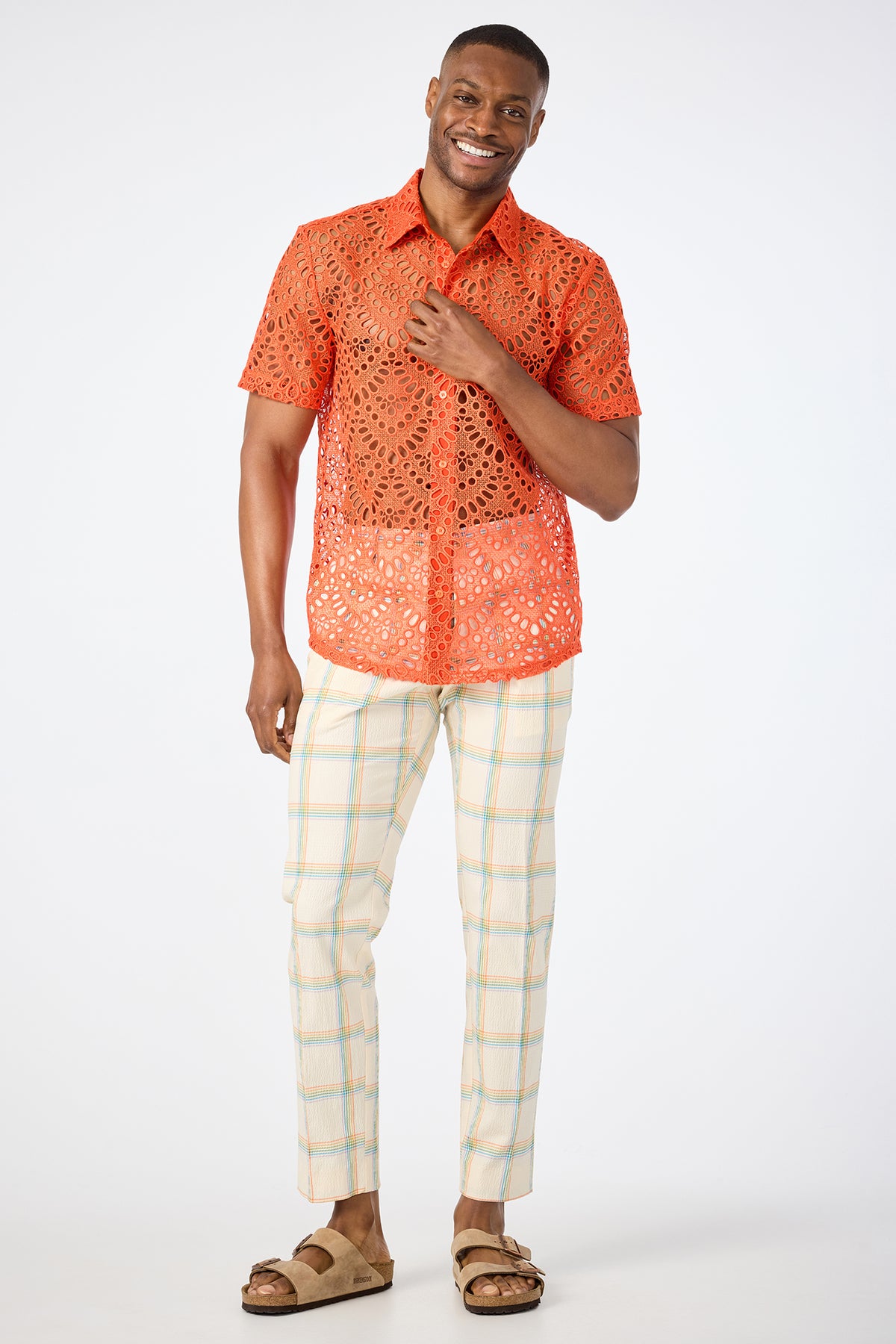A man smiles while wearing the Long Beach Short-Sleeve Shirt in bright orange, light plaid pants, and tan sandals, standing against a plain white background.