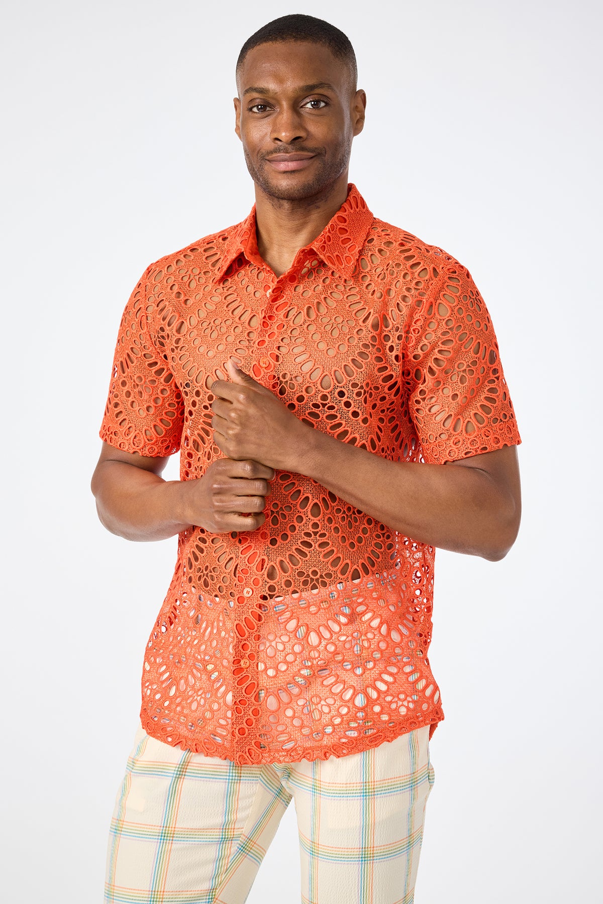 A man smiles softly and clasps his hands together while wearing the Long Beach Short-Sleeve Shirt in Marseille Mandarin with circular lace patterns and light plaid pants, standing against a plain background.