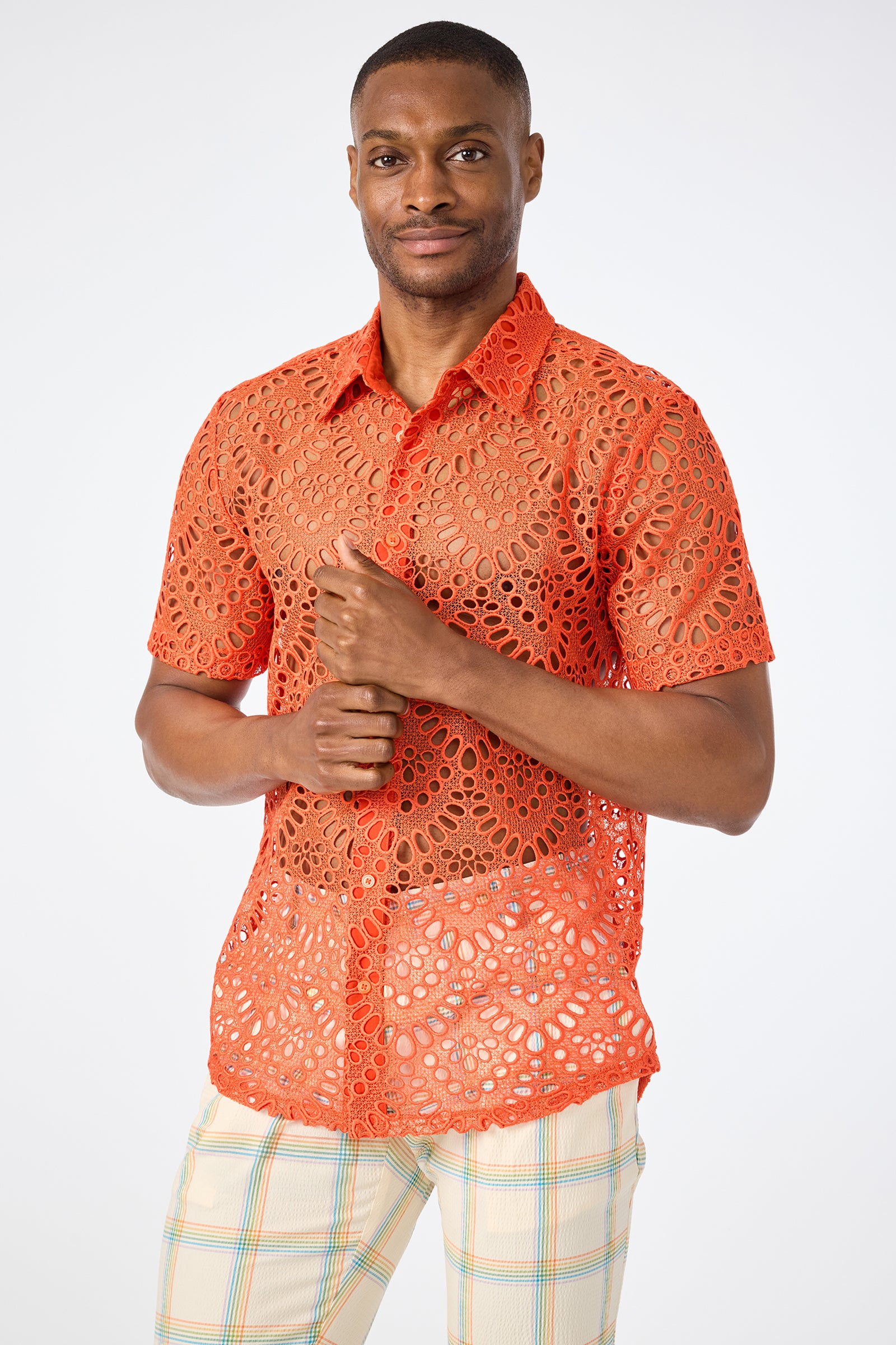 A man smiles softly and clasps his hands together while wearing the Long Beach Short-Sleeve Shirt in Marseille Mandarin with circular lace patterns and light plaid pants, standing against a plain background.