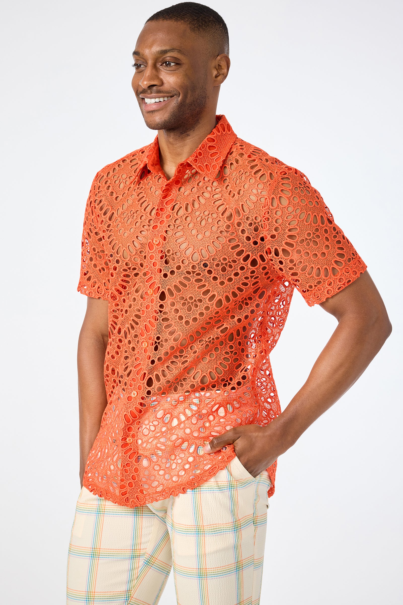 A smiling man with one hand in his pocket wears the Long Beach Short-Sleeve Shirt, a bright orange lace top with circular cutouts, paired with light plaid pants against a plain white background.