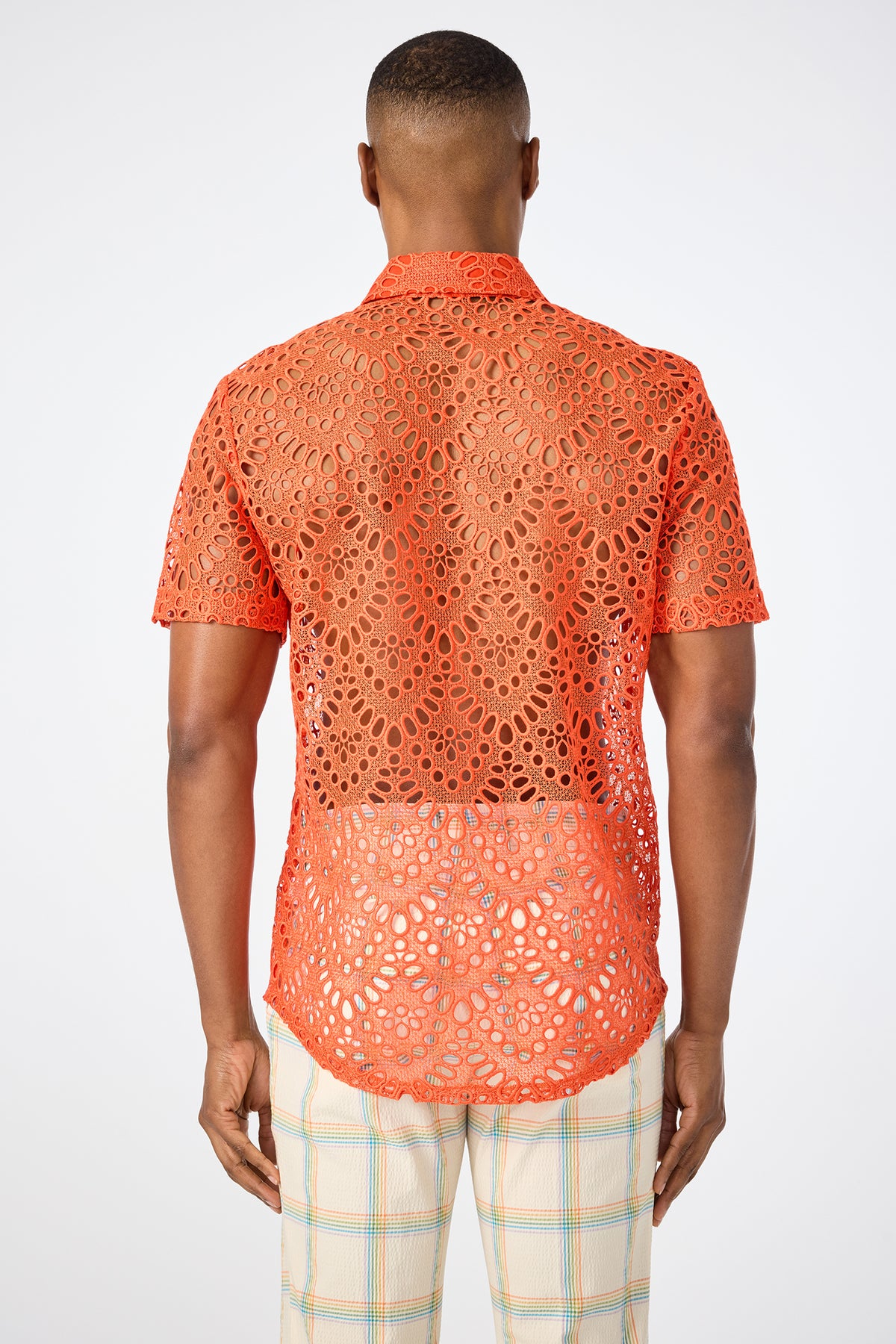 A person stands facing away, wearing the Long Beach Short-Sleeve Shirt in bright orange with a see-through lace pattern and light-colored plaid pants, set against a plain white background.