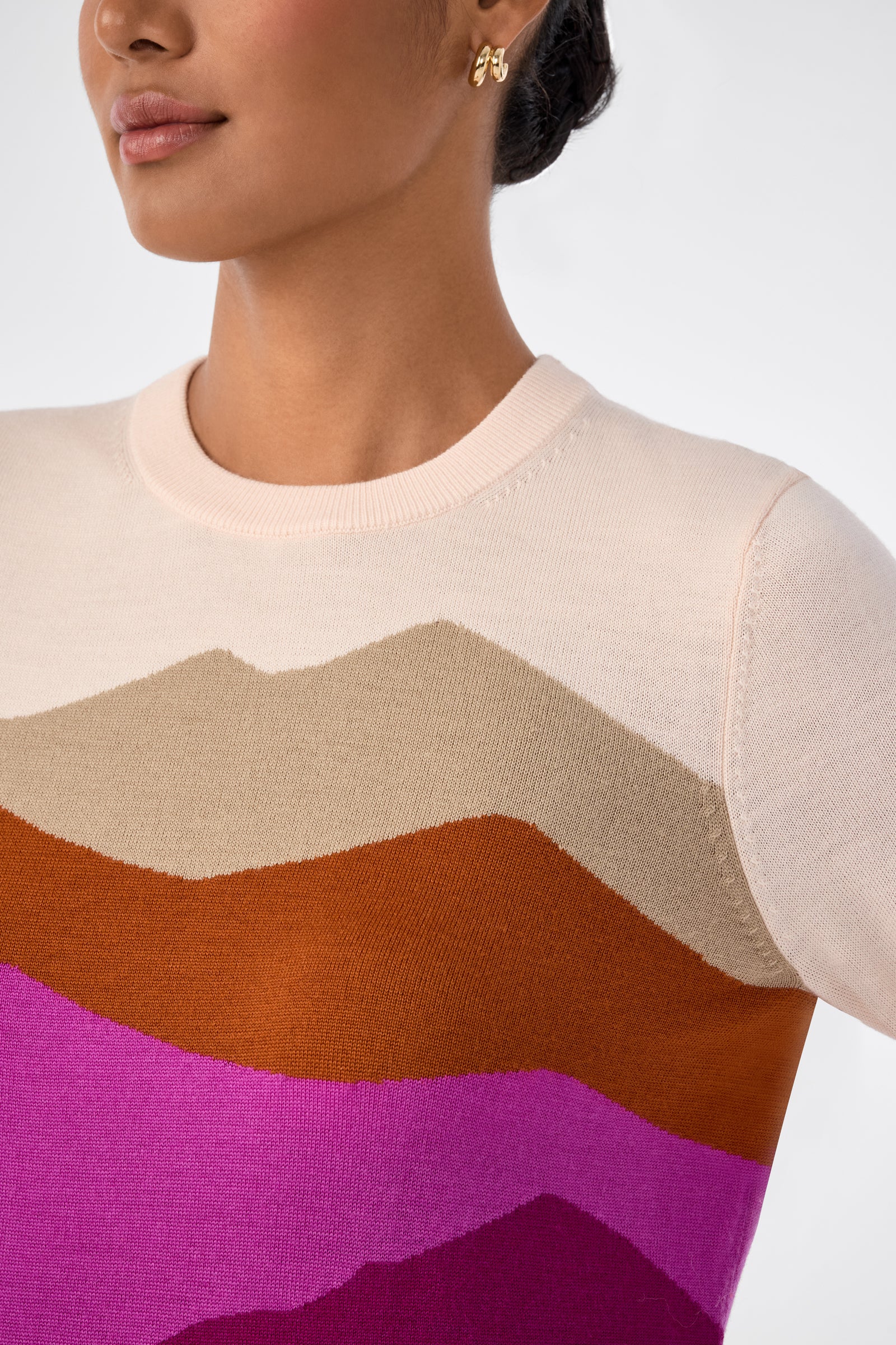 A woman wears the Jacinto Crewneck, a cream-colored sweater with bold wavy stripes in tan, brown, orange, and pink. She has gold earrings and her hair is styled back. The image is a chest-up shot against a plain background.