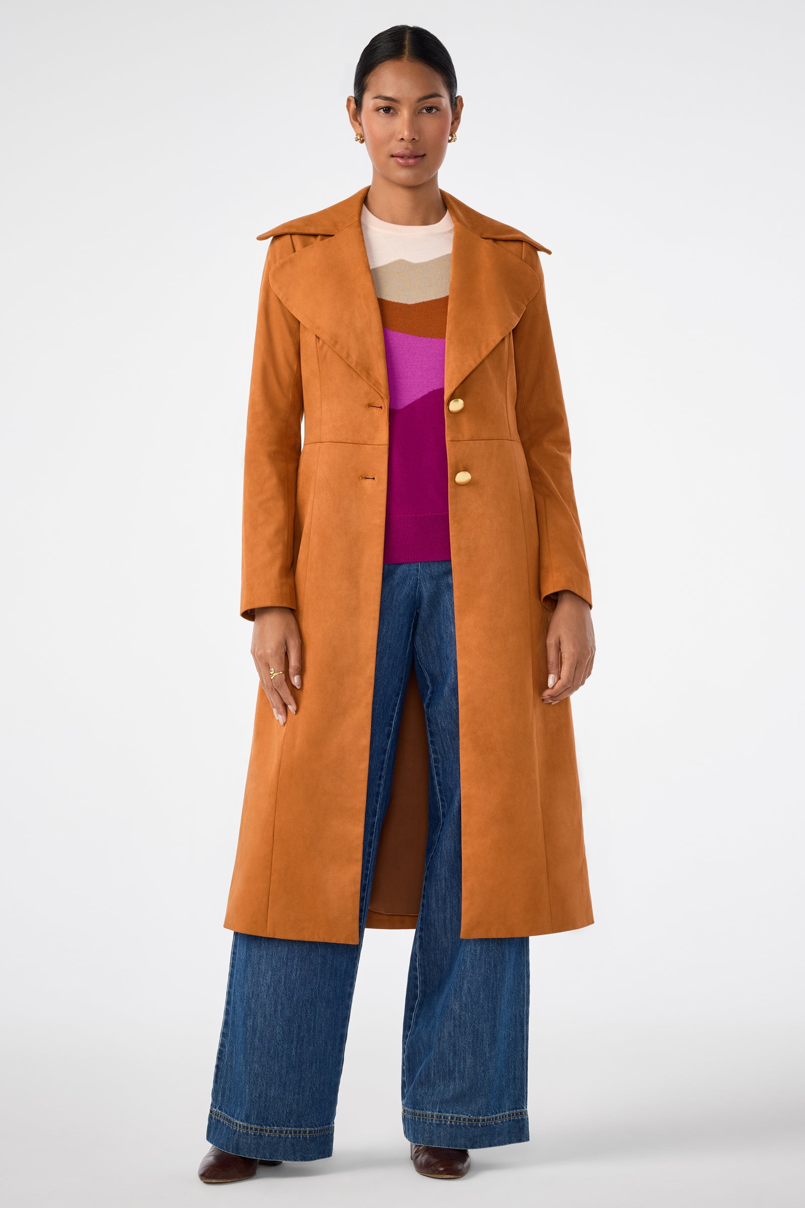 A person stands facing forward in a Jacinto Crewneck, an orange-brown long coat, wide-leg blue jeans, and brown shoes against a plain white background.