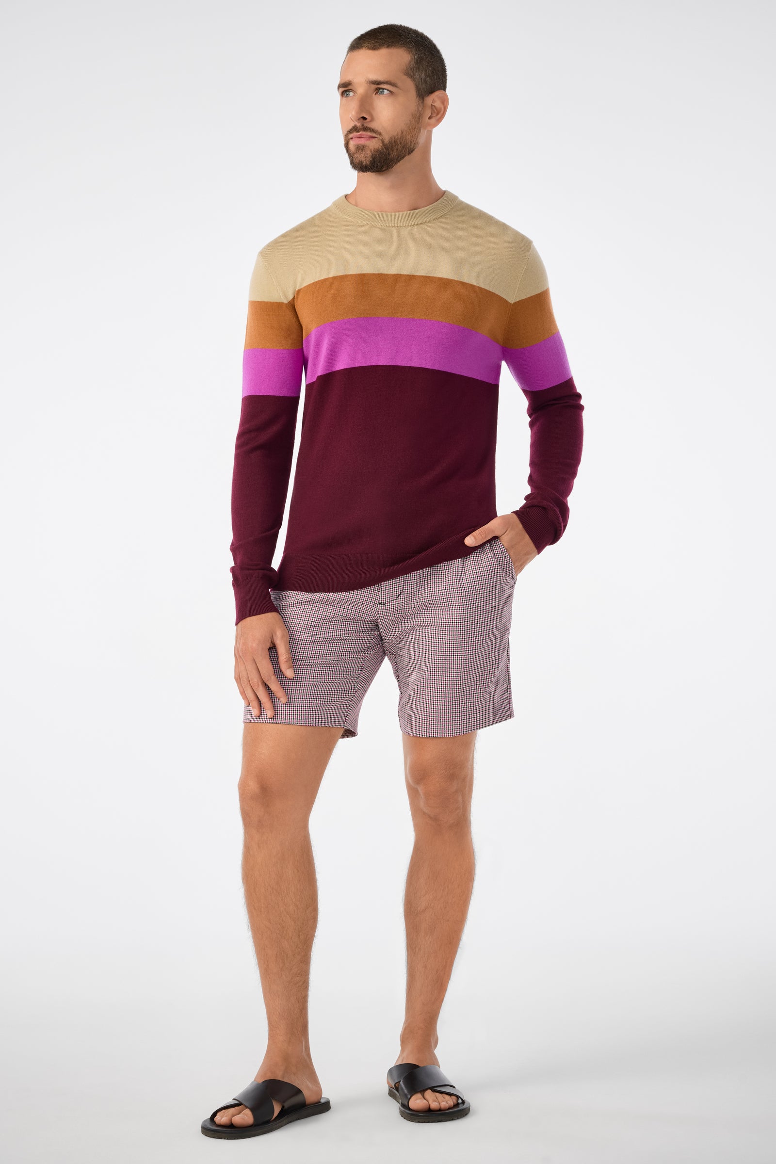 A man wears the Romero Sweater, featuring tan, mustard, purple, and burgundy color-block stripes, paired with plaid shorts and black sandals against a plain white background.