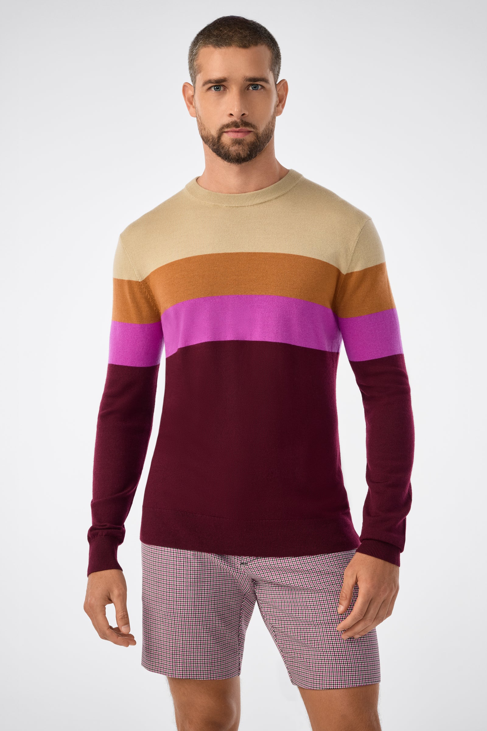 A man with short hair and a beard wears the Romero Sweater, featuring beige, mustard, purple, and burgundy color-block stripes, paired with checked shorts. He stands facing forward against a plain light background.
