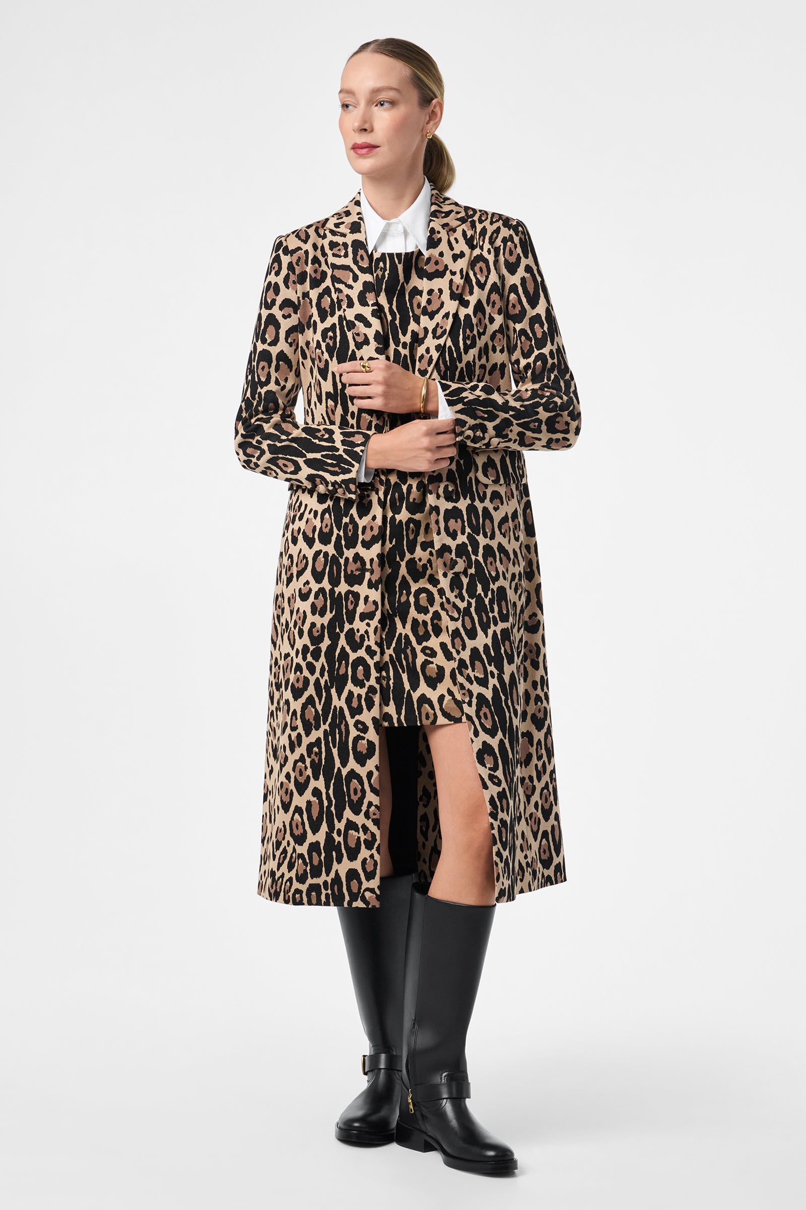 A woman models the Playbill Coat—long, leopard-print style—over a white collared shirt and black knee-high boots, standing against a white background with her blonde hair pulled back and holding the coat closed.