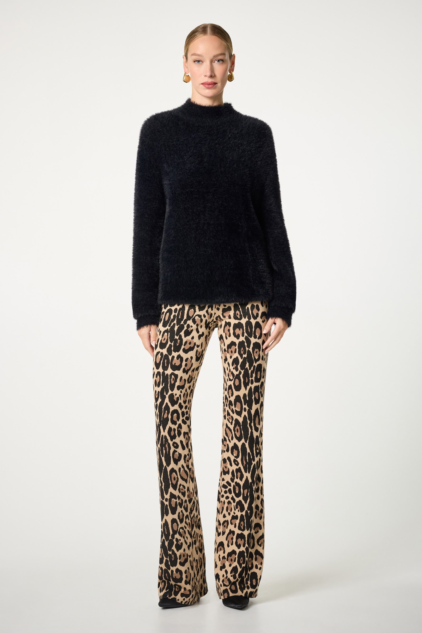 A woman stands against a plain background wearing the Bazaar Mock Neck Sweater, gold hoop earrings, high-waisted flared leopard print pants, and black platform shoes.